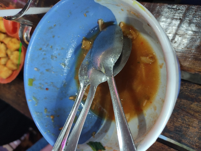The image captures a close-up, overhead view of a blue bowl with a white rim, mostly empty but containing remnants of a reddish-orange sauce and a few small food particles. Two silver-colored metal spoons rest inside the bowl, partially covered by the remaining liquid.
To the upper left, a portion of a clear glass or cup is visible. Further left, part of a red or orange basket or container can be seen, filled with numerous small, golden-brown, cuboid-shaped fried items, likely fried tofu or tempeh. All items are situated on a dark wooden table surface, which shows visible grain.
The scene depicts the aftermath of a meal or snack, indicated by the nearly empty bowl with leftovers. No people are visible, nor is any text. The setting is a casual environment, consistent with a mealtime in Kecamatan Pandaan, Indonesia. The lighting is ambient, without strong highlights or shadows.