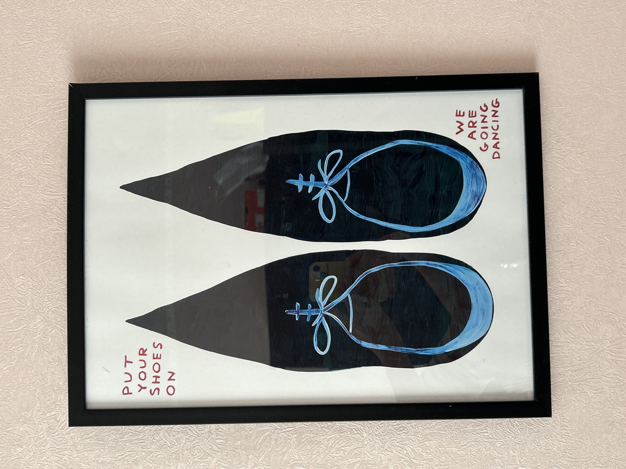 The image shows a framed artwork hanging on a wall with textured pink wallpaper. The artwork features a white background with two black, elongated shapes resembling pointe shoes. Each shoe has a blue outline and a blue ribbon tied in a bow with a cross-like embellishment above it. Red text on the left side of the artwork reads "PUT YOUR SHOES ON," and red text on the right side reads "WE ARE GOING DANCING." The scene is an indoor setting, possibly a room in a residential or commercial building. There is no indication of the specific location within the city of Moscow, Russia.