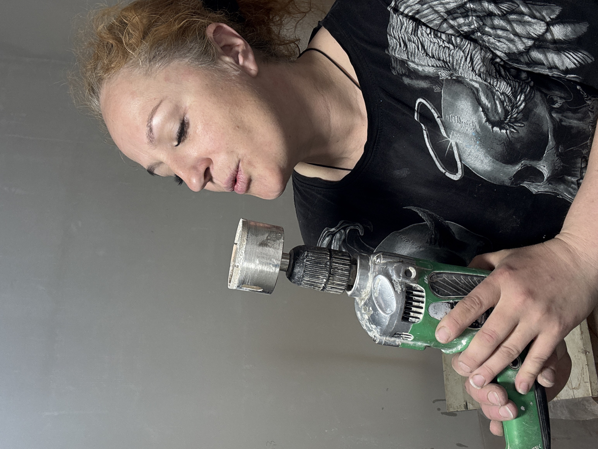 A woman with light skin and reddish-blonde hair, styled in a loose updo, is shown from a slightly high, tilted angle holding a green and silver Hitachi power drill with a hole saw attachment. She is looking down at the drill with her eyes closed or partially closed. She is wearing a black t-shirt with a silvery, graphic design on the front. The background appears to be a plain, light-gray wall. The image does not provide any visual information about the location in the city of Chaikovsky, Russia.