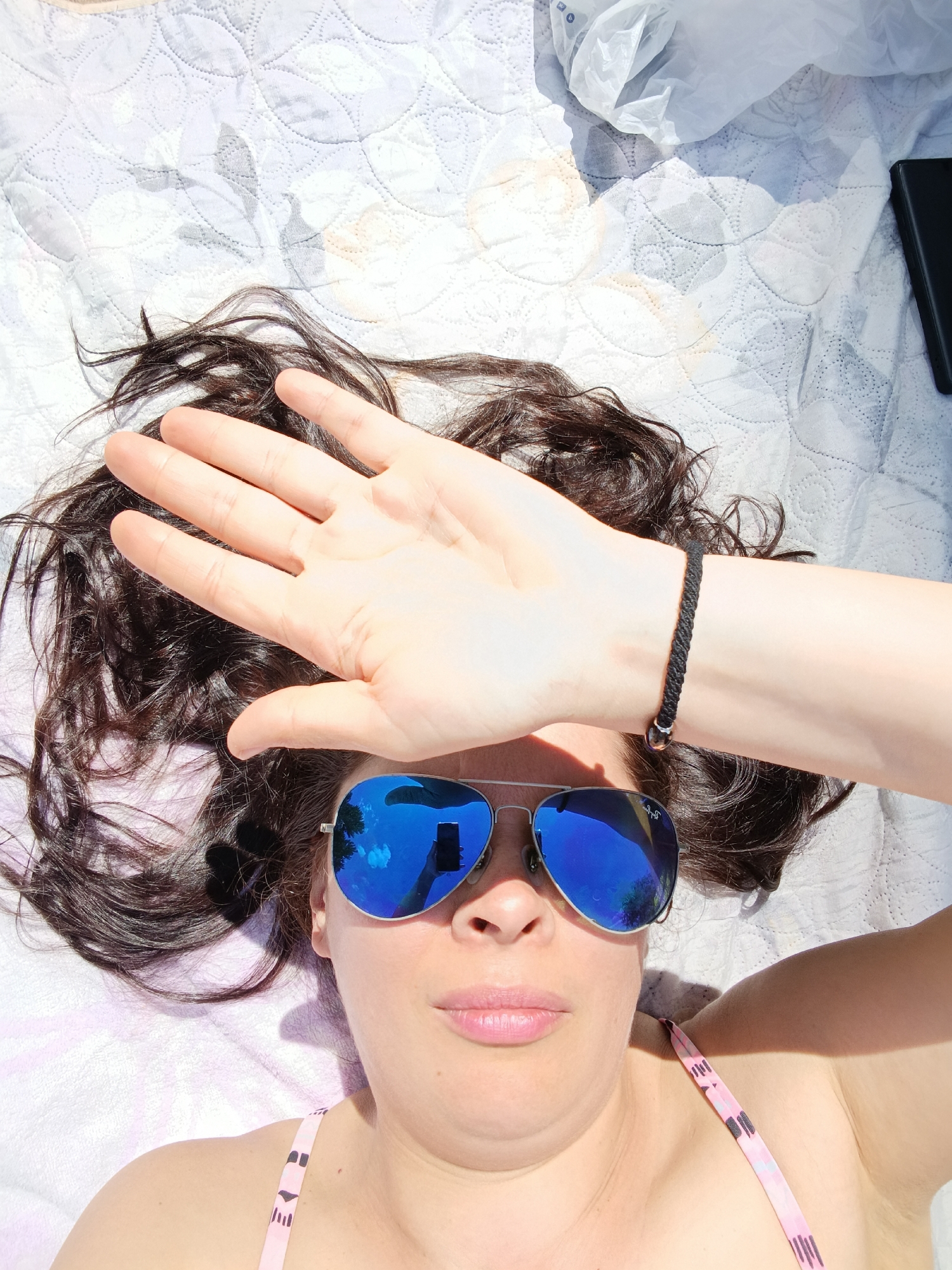A woman is lying on a blanket outdoors on a sunny day, covering her face with her right hand. She is wearing aviator sunglasses with blue mirrored lenses, and a pink bikini top. Her dark hair is spread around her head. A black braided bracelet with a silver ring is on her left wrist. The blanket is white with a patterned design and appears to be on a surface that is also white and textured. There is a dark rectangular object to the upper right of the woman. There are also crumpled white plastic items to the upper right. The scene is illuminated by bright sunlight, casting shadows on the blanket. There is no indication of the city of Saratov, Russia.