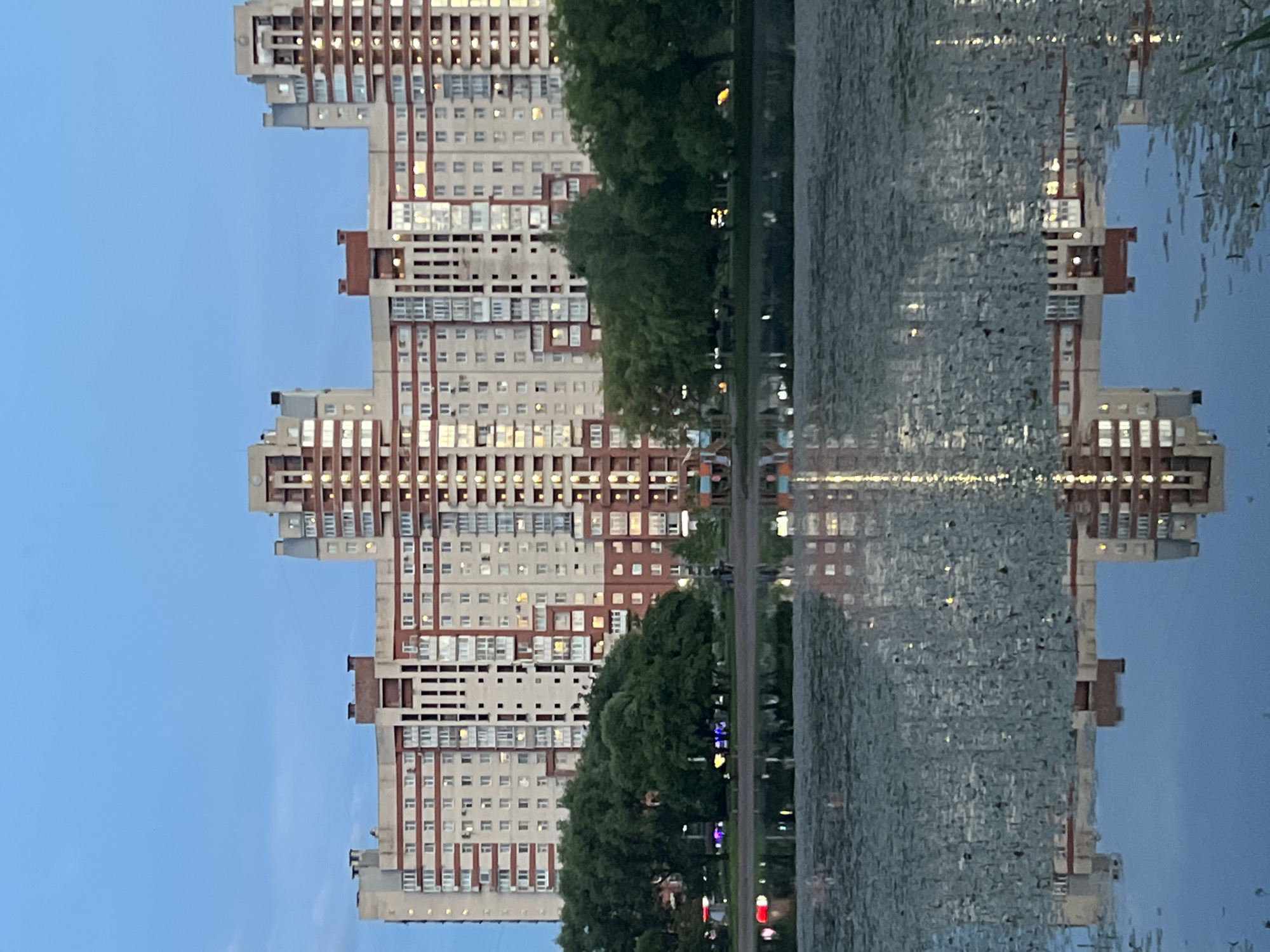 In Irinovka, Russia, a reflection of a large, multi-story apartment complex is visible in a body of water. The apartment complex features a light-colored facade with red brick accents and numerous windows, some of which are illuminated. The water in the foreground is partially covered with aquatic plants, and the sky above is a clear, light blue. Trees line the edge of the water, and a park area with playground equipment is discernible behind them. The overall scene depicts a tranquil evening view of a residential area.