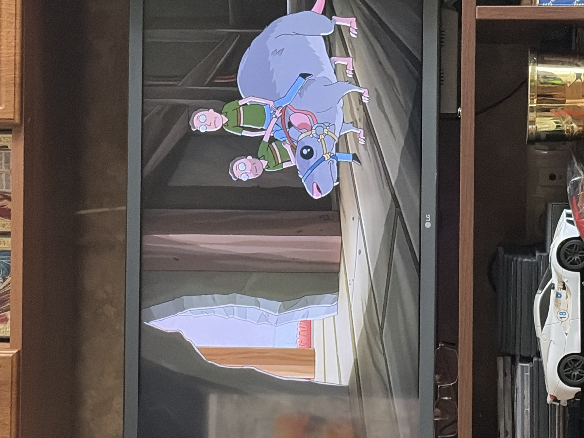 In an animated scene depicting a fantasy or science fiction environment, two characters resembling humans, wearing green shirts with striped accents and blue pants, are positioned on the back of a large, light-gray rodent. The character in front is seated, appearing to steer the rodent with reins. The rodent has a long tail, large ears, and is wearing a blue and red halter. The characters and rodent are on a wooden-planked floor, with a large, cave-like opening to the left that reveals a brightly lit area with red structures in the distance. The background consists of wooden walls and structures. The image is a photograph of a television screen displaying this scene.