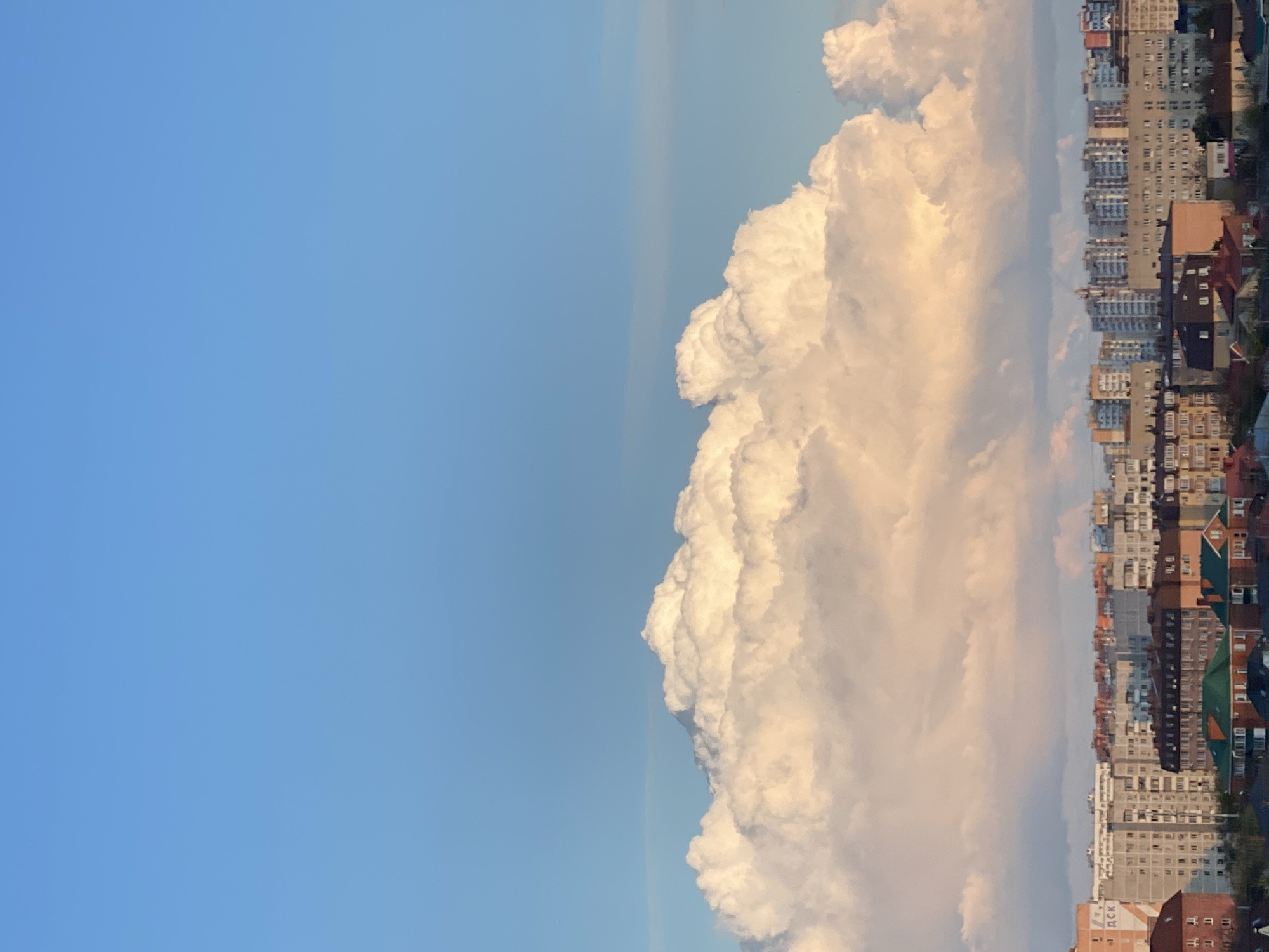 The image depicts a cityscape, likely of Unknown, Unknown, with a large cloud formation dominating the central view. To the left is a clear blue sky. The cloud is white with shadows and sunlit areas giving it a three-dimensional appearance. The city is comprised of numerous apartment buildings, each appearing multi-storied. The buildings vary in color, including shades of gray, tan, and brick red. The rooftops of some buildings are visible, some flat and some peaked. The scene is lit with natural daylight.