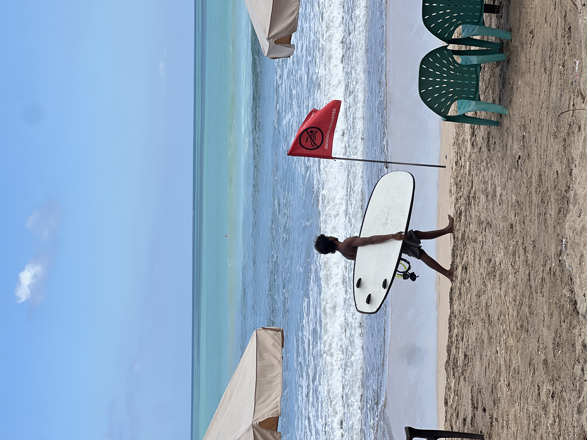 A man walks along the beach in Kuta, Indonesia, carrying a white surfboard. He is wearing dark shorts and his back is to the camera. To his right, a red flag with a white circle and diagonal line through a swimmer icon, and the words "SWIMMING PROHIBITED" in white text, is planted in the sand. Two green plastic chairs are visible to the right of the flag. In the background, the turquoise ocean meets the blue sky. Two beige umbrellas are visible on the left and right sides of the image, providing shade. The sand is light brown and textured.