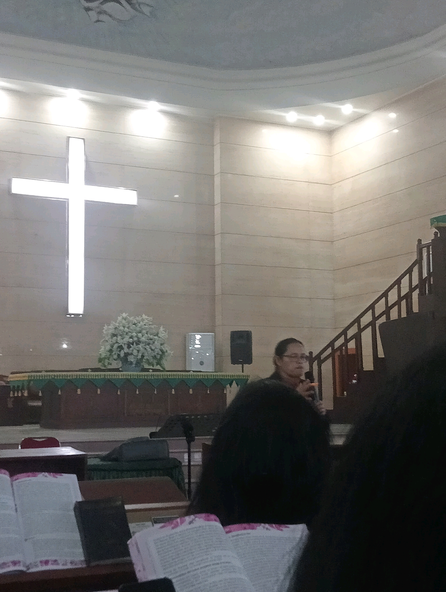 The image shows the interior of a church with a large illuminated cross on the wall. A woman is standing and speaking into a microphone, possibly giving a sermon or a speech. She is wearing glasses and a dark top. In the foreground, there are open books, likely bibles. The church has wooden elements, including a pulpit or stage area with a decorative cloth and a staircase. The overall lighting is warm and diffused. Without specific visual cues or information beyond what is present in the image itself, it is not possible to determine the location within the city of Jambi, Indonesia.