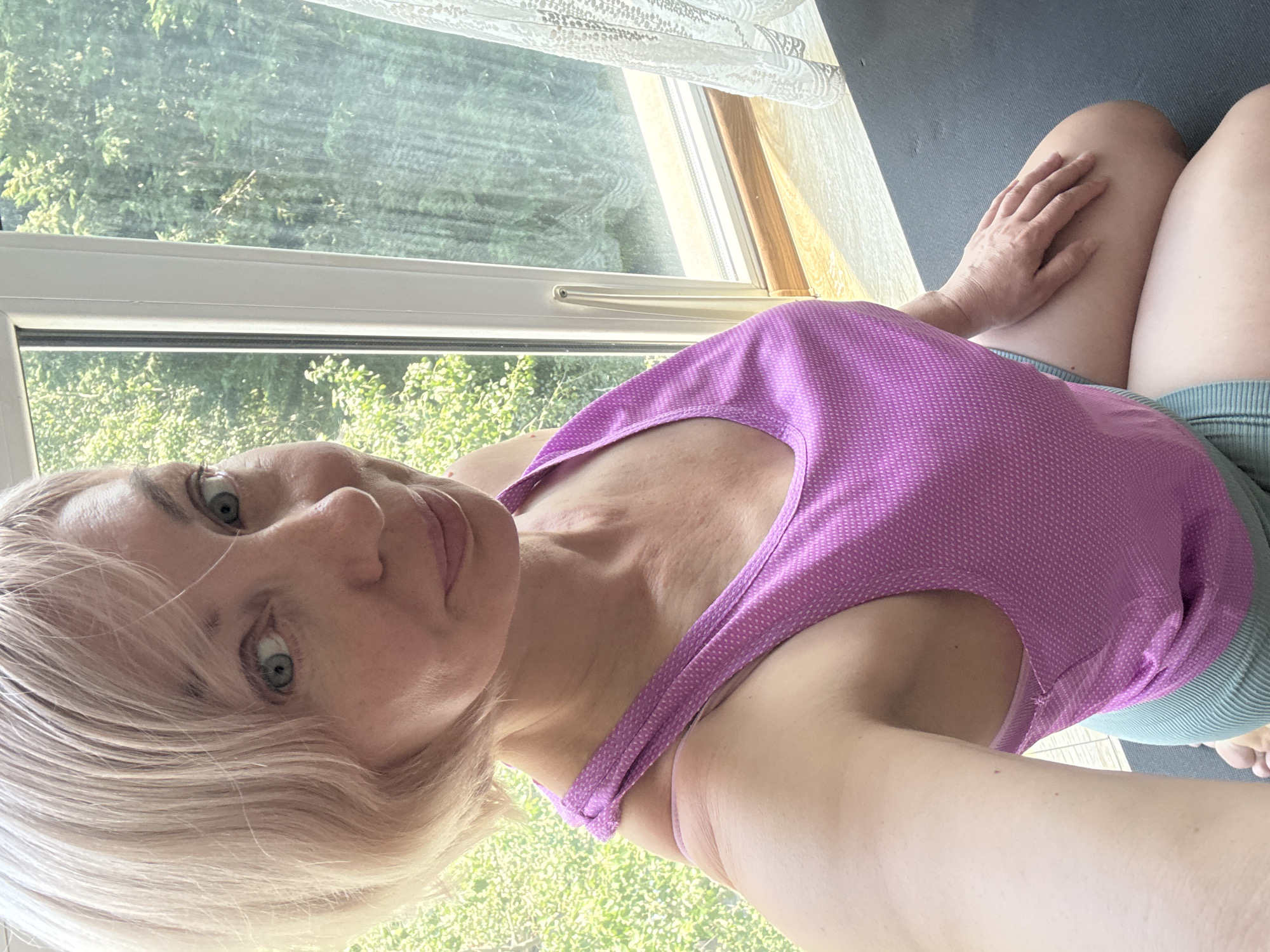 An indoor selfie captures a light-skinned female, appearing middle-aged, with short, light blonde hair and blue-green eyes, looking directly at the camera with a subtle smile. She is wearing a magenta athletic tank top with a fine textured pattern and light green ribbed shorts, kneeling on a dark grey yoga mat. The scene is located in a room in Sochi, Russia, with the subject positioned in front of a white-framed window. Outside the window, dense green foliage, consistent with a subtropical climate, is visible under bright daylight. A wooden windowsill extends below the window, and a white, patterned lace curtain is partially visible on the right side of the frame. Her right hand rests on her right thigh.