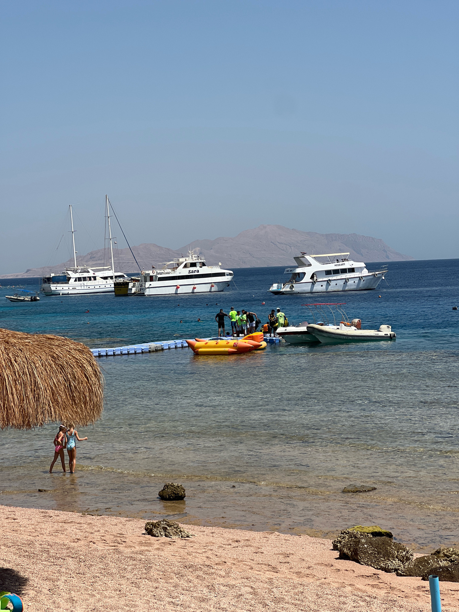 The image depicts a scene on the coast of Sharm El Sheikh, Egypt, under a clear, bright sky. In the foreground, a sandy beach with scattered rocks is visible, with two young girls in swimsuits standing at the edge of the shallow, clear water. A thatched umbrella shades a portion of the beach.
Further out in the turquoise sea, several boats are anchored. Two large white yachts, one with "Sara" printed on its side, are prominent. A sailboat with tall masts is also present. A floating dock with several people, some wearing life vests, is connected to the shore by a blue floating walkway. A yellow inflatable watercraft and two smaller motorboats are docked or moored near the floating structure. In the background, rugged, mountainous terrain is visible, characteristic of the Sinai Peninsula. The water is calm, with gentle ripples on the surface. Several other individuals can be seen swimming or relaxing in the water. The overall impression is a vibrant tourist destination, likely a beach resort.