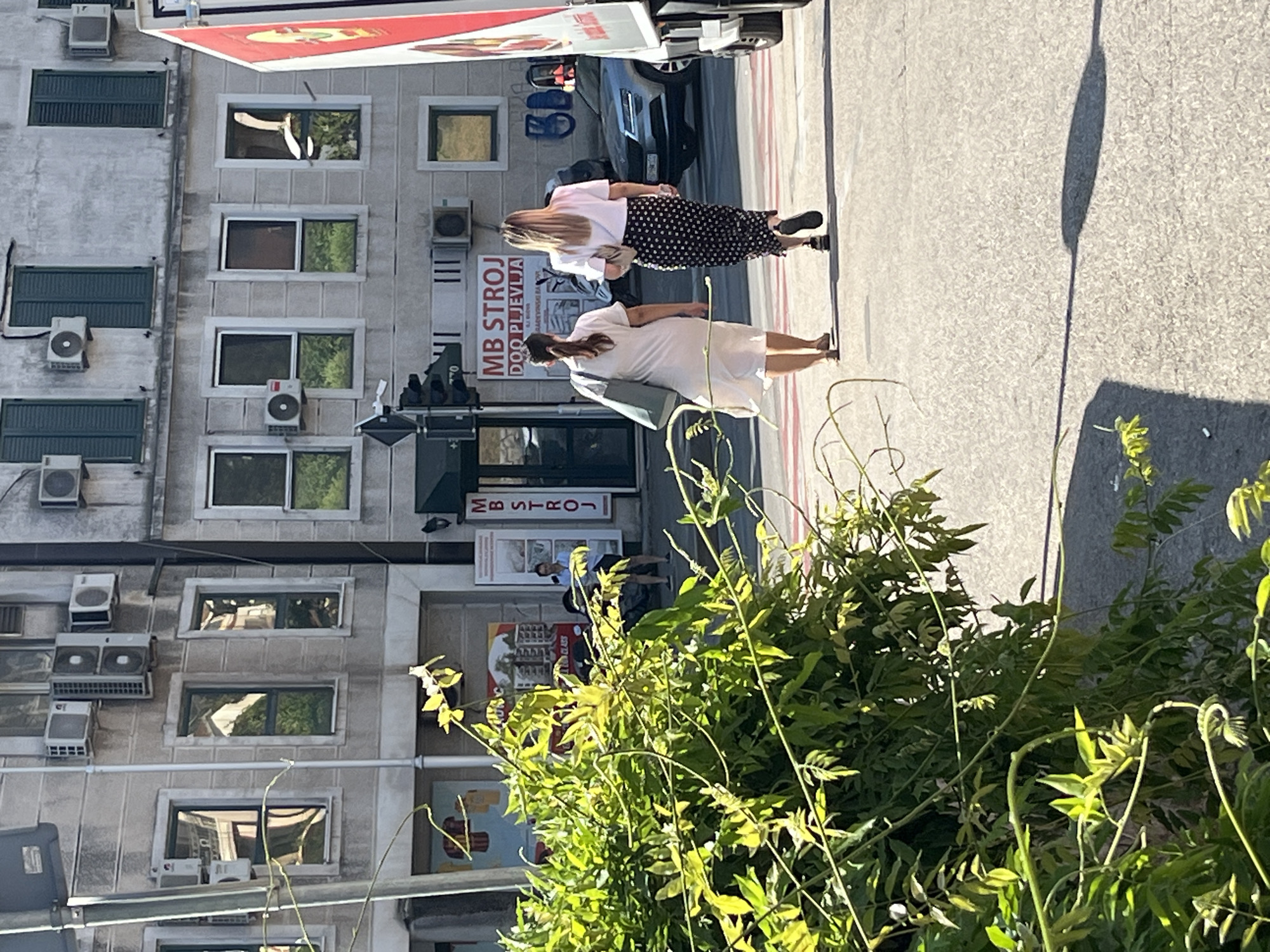 The image depicts a street scene in Budva, Montenegro, on a sunny day. In the foreground, lush green foliage partially obscures the view. Two women are walking away from the camera on a paved street, with their backs to the viewer. The woman on the left wears a white dress and carries a light-colored bag. The woman on the right is dressed in a white t-shirt and a black skirt with white polka dots, and she carries a beige bag. Behind them, a silver car is parked. The background features a multi-story building with windows and air conditioning units. A sign on the building reads "MB STROJ DOO PLJEVLJA". A portion of a truck is visible on the right side of the frame. The overall impression is of an ordinary urban environment.