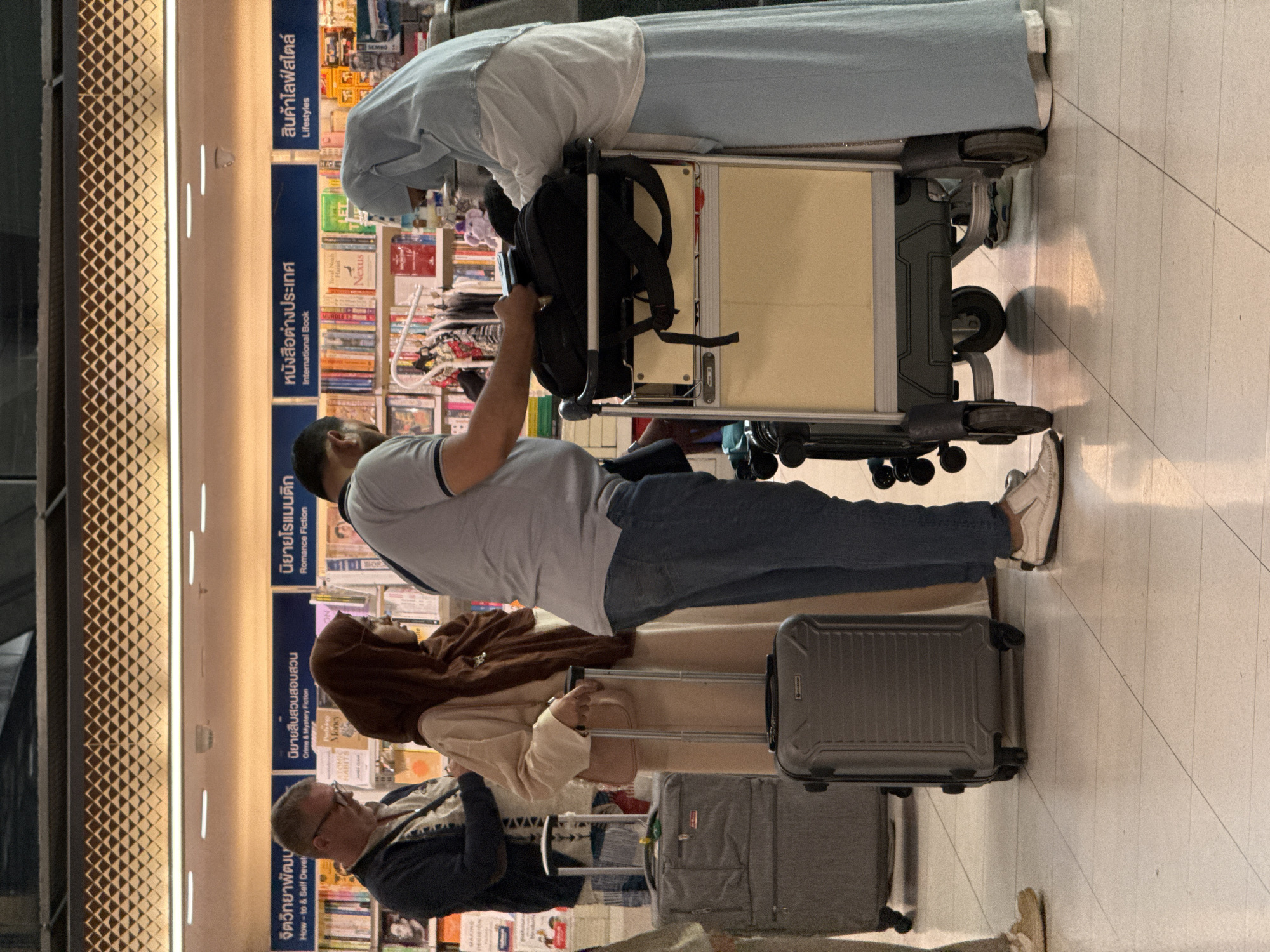 In the city of Nong Prue Subdistrict, Thailand, a group of individuals is gathered in a bookstore, likely an airport terminal given the presence of luggage. The scene is illuminated by overhead lighting, casting reflections on the tiled floor. Three individuals are prominently visible. To the left, a man wearing glasses and a dark jacket over a patterned shirt stands facing a bookshelf. Next to him, a woman wearing a hijab and a long, light-colored garment is holding the handle of a rolling suitcase. Her right hand is on the handle, and her left hand is resting on a pink purse. To the right of the woman, a person in a light blue head covering and a long blue garment is interacting with a luggage cart. The cart has a yellow compartment and a black bag on top. Another person, partially visible on the far right, appears to be wearing a similar blue garment. A third rolling suitcase, gray and upright, stands to the left of the woman in the light garment. Bookshelves filled with various titles line the background, with signage in Thai indicating sections such as "How-to & Self Development," "Romance Fiction," "International Book," and "Lifestyles." The overall impression is one of travel and commerce.