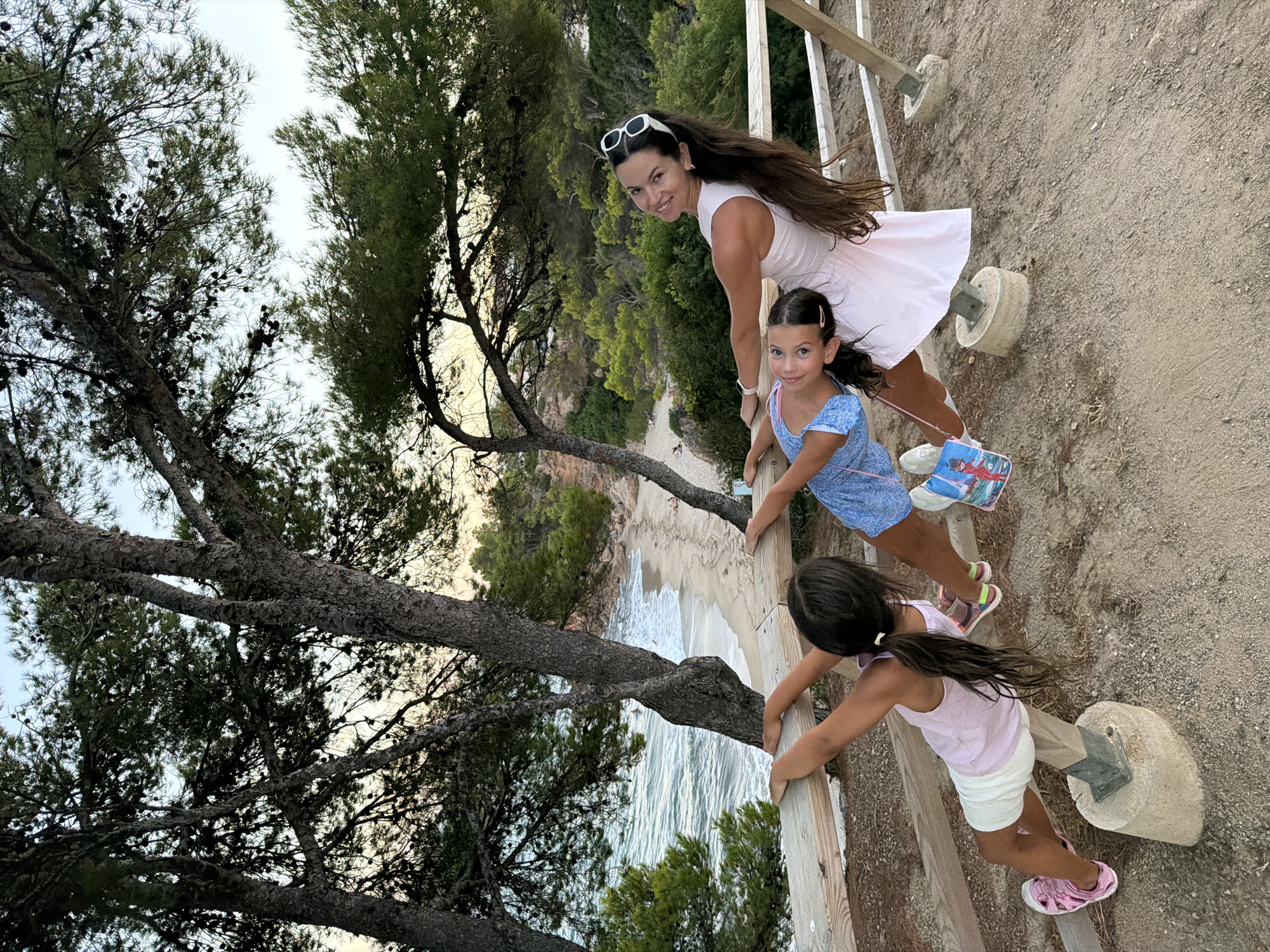 This image depicts a woman and two young girls on a wooden railing overlooking a beach and the Mediterranean Sea in Mont-roig del Camp, Spain. The woman, wearing a white dress and white sunglasses, smiles at the camera while leaning on the railing. She has long dark hair and is positioned on the right side of the frame. To her left, the two girls are also interacting with the railing. The girl on the left, wearing a light pink tank top and white shorts, is leaning back over the railing with her legs bent. The girl on the right, wearing a blue patterned dress, is leaning forward. Both girls have dark hair and are positioned towards the left side of the frame. The background features a sandy beach with gentle waves, lush green trees, and a clear sky. The scene appears to be set in the late afternoon or early evening.
