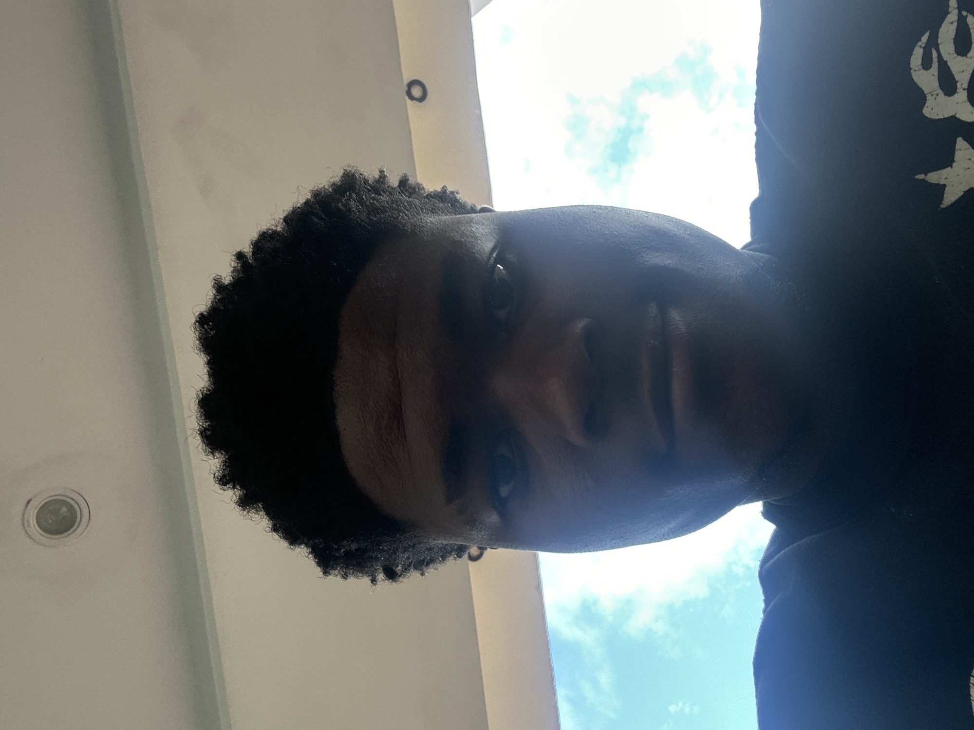 The image is a close-up, low-angle selfie of a young Black male with dark, curly hair, looking directly at the camera. He is wearing a dark t-shirt, visible at the bottom of the frame. The background is a bright sky with white clouds, suggesting an outdoor setting. The ceiling above him is visible at the top of the image, with a circular light fixture. The scene is likely from Ihiagwa, Nigeria, given the user's prompt.