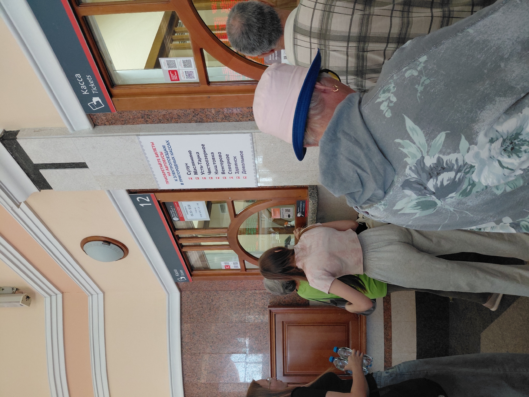 The image shows a ticket counter at what appears to be a train or bus station. Several people are visible in line, waiting to purchase tickets. The ticket counter has a "Kacca Tickets" sign and a large window through which transactions are conducted. There are informational posters displayed on the wall, some of which are in Russian. The environment is indoors, with a tiled floor and a patterned wall. The lighting suggests it is daytime.