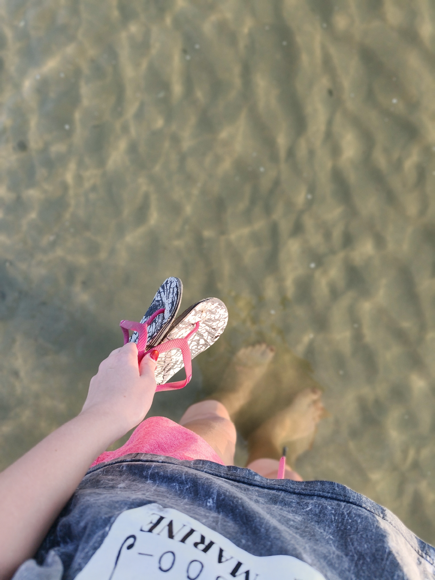A person's hand, adorned with a red nail polish, holds a pair of pink and black patterned flip-flops over shallow, clear water. The person's legs and feet are submerged in the water, wearing bright pink shorts and a dark grey t-shirt with white text. The scene appears to be a beach or shallow body of water, potentially in Đà Nẵng, Vietnam, given the context of the request. The water has a sandy bottom, visible through the rippling surface, creating patterns of light and shadow.