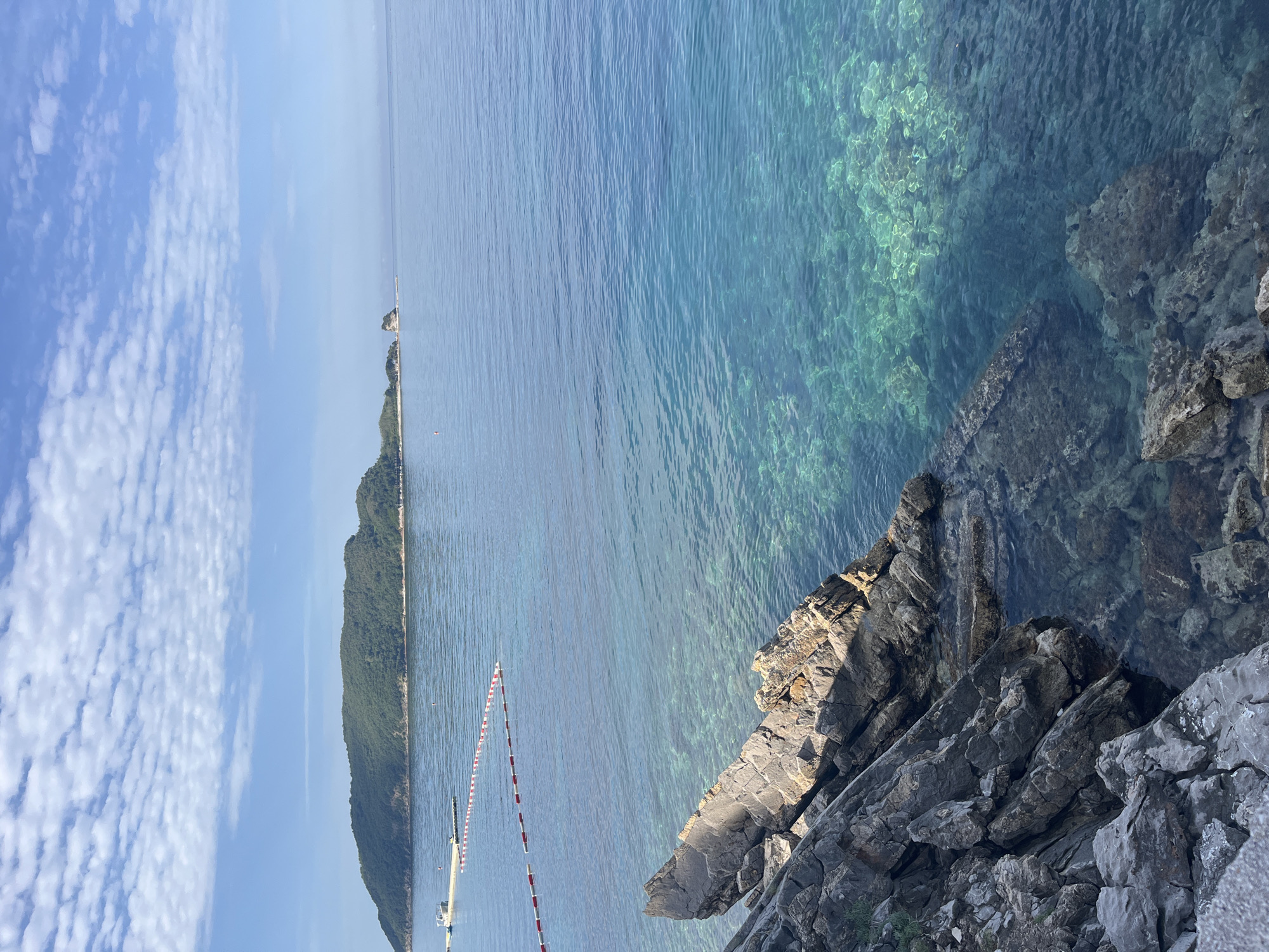 The image depicts a coastal scene in Boreti, Montenegro. The foreground features rocky cliffs sloping into a body of clear, turquoise water. In the midground, there is a long, narrow peninsula covered in green vegetation, extending out into the water. A row of red and white buoys marks a line along the peninsula. In the background, the sky is blue with scattered white clouds. The water reflects light, and the scene is generally bright and well-lit.