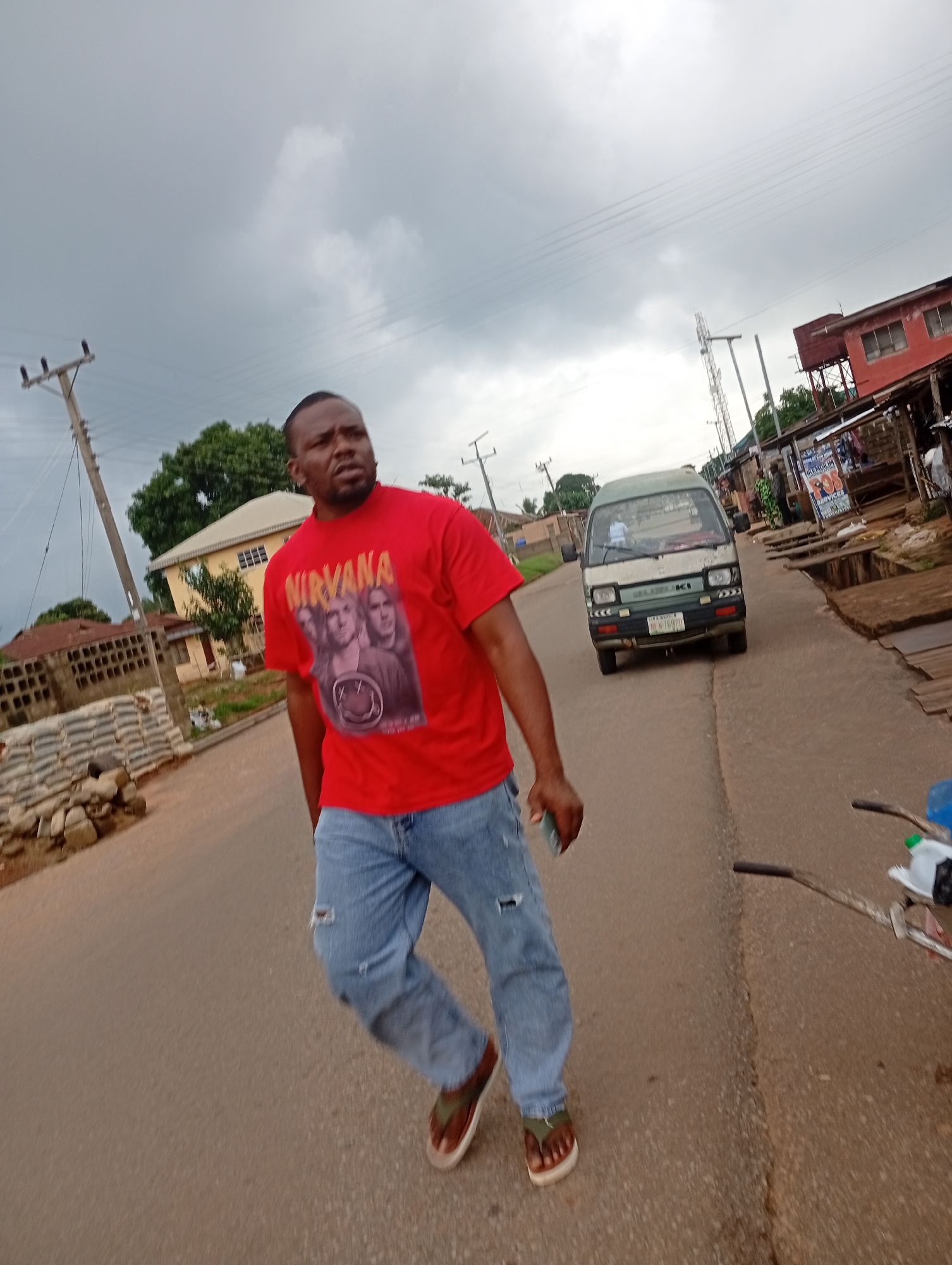 A man wearing a red Nirvana t-shirt and blue ripped jeans stands on an asphalt road in Nsugbe, Nigeria. He is holding a mobile phone in his right hand. The man is looking towards the right side of the frame. Behind him, a white van with a green stripe is driving down the road. In the background, there are buildings, including a red-colored building with an orange roof, and a white house with a sloped roof. There are also power lines and trees visible. The sky is cloudy. The scene appears to be a typical street in a Nigerian town.
