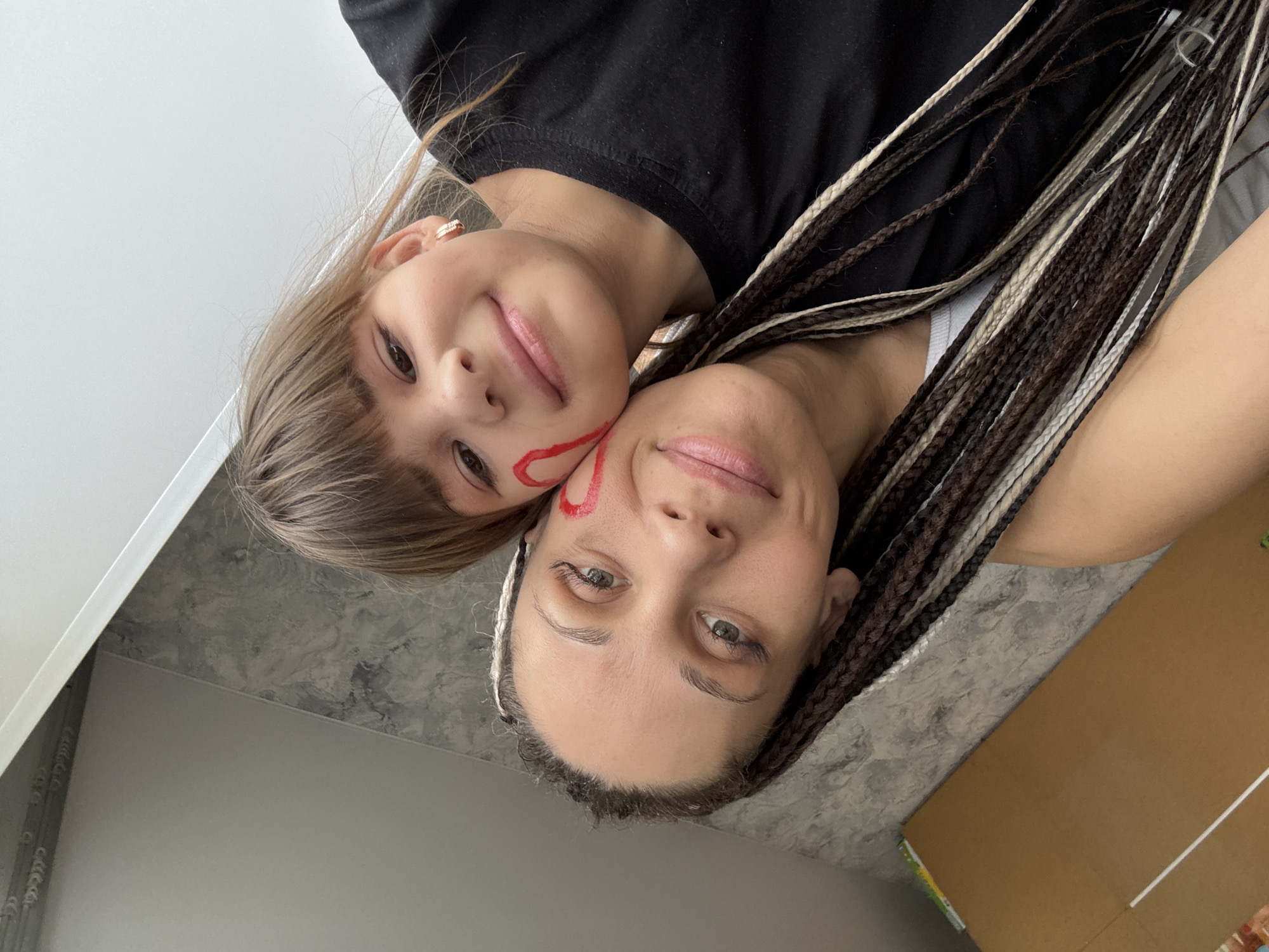 A selfie photograph shows a woman and a child posing closely together. The child is smaller, with straight blonde hair, wearing a black shirt, and has a red, heart-shaped marking on her face. The woman has dark hair styled in braids with blonde highlights, and a similar red marking on her cheek. Both are looking directly at the camera and appear to be smiling.
The background includes a white wall and a grey, marble-patterned surface, possibly a countertop or floor. A cardboard box is also partially visible.
It is not possible to ascertain the location in the city of Rusanovo, Russia, from the image alone.