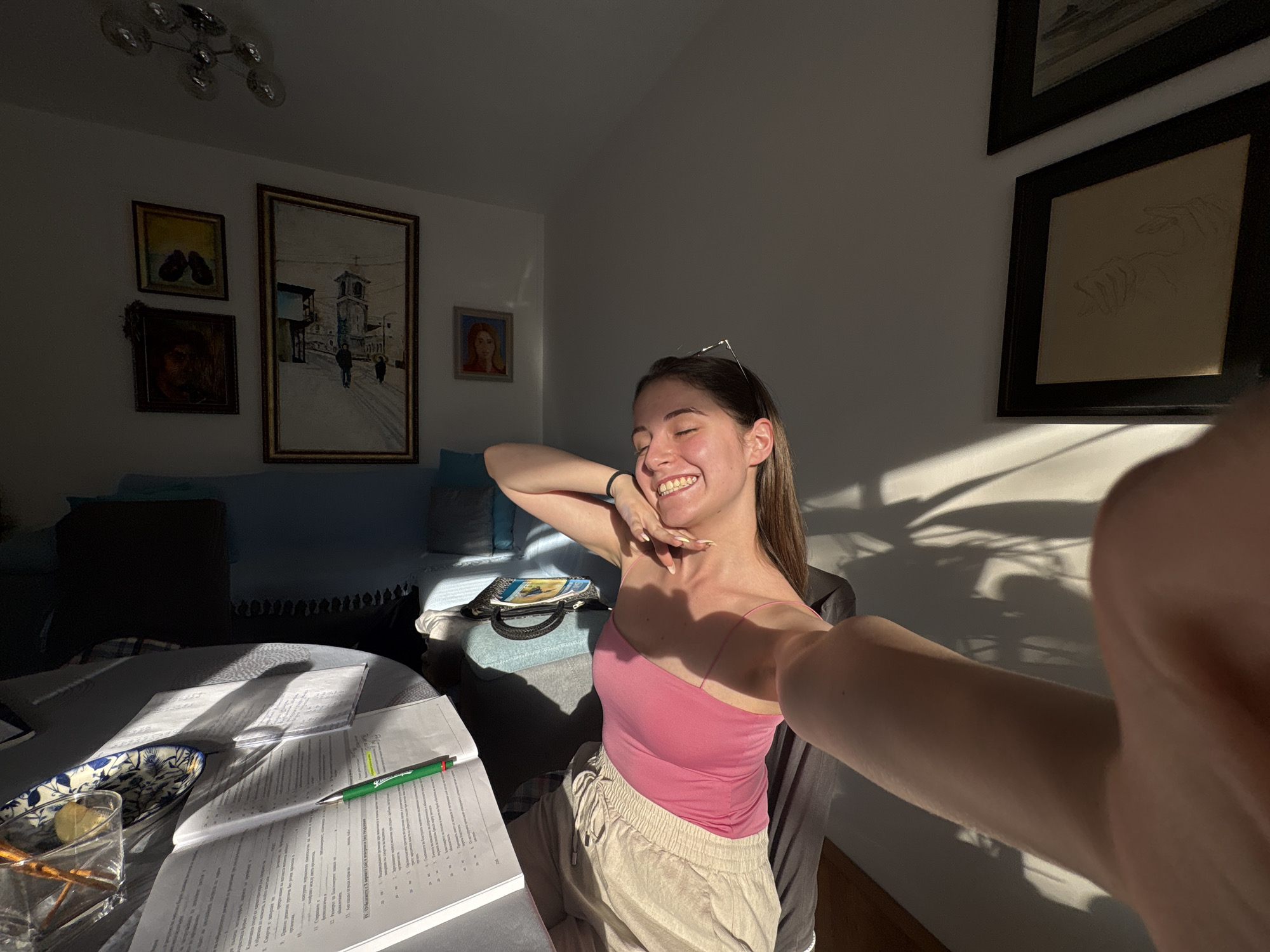 A young woman with long brown hair, wearing a pink tank top and beige shorts, is taking a selfie in a sunlit room. She has her eyes closed and is smiling, with her left arm raised behind her head and her right hand near her face. Her glasses are pushed up onto her head.
The scene appears to be a home office or study area. A round table in the foreground is covered with open books, papers, a pen, a glass of water with straws, and a small plate with a slice of lemon. The sunlight streams in from the right, casting strong shadows across the table and onto the wall.
In the background, a blue sofa is visible, with several framed pictures hanging on the wall above it. One painting depicts a European street scene with a church tower, another shows a portrait of a woman, and a third is a sketch of hands. A modern chandelier hangs from the ceiling. The overall impression is one of a relaxed and bright afternoon, possibly during study time. The visible text on the papers appears to be in Bulgarian.