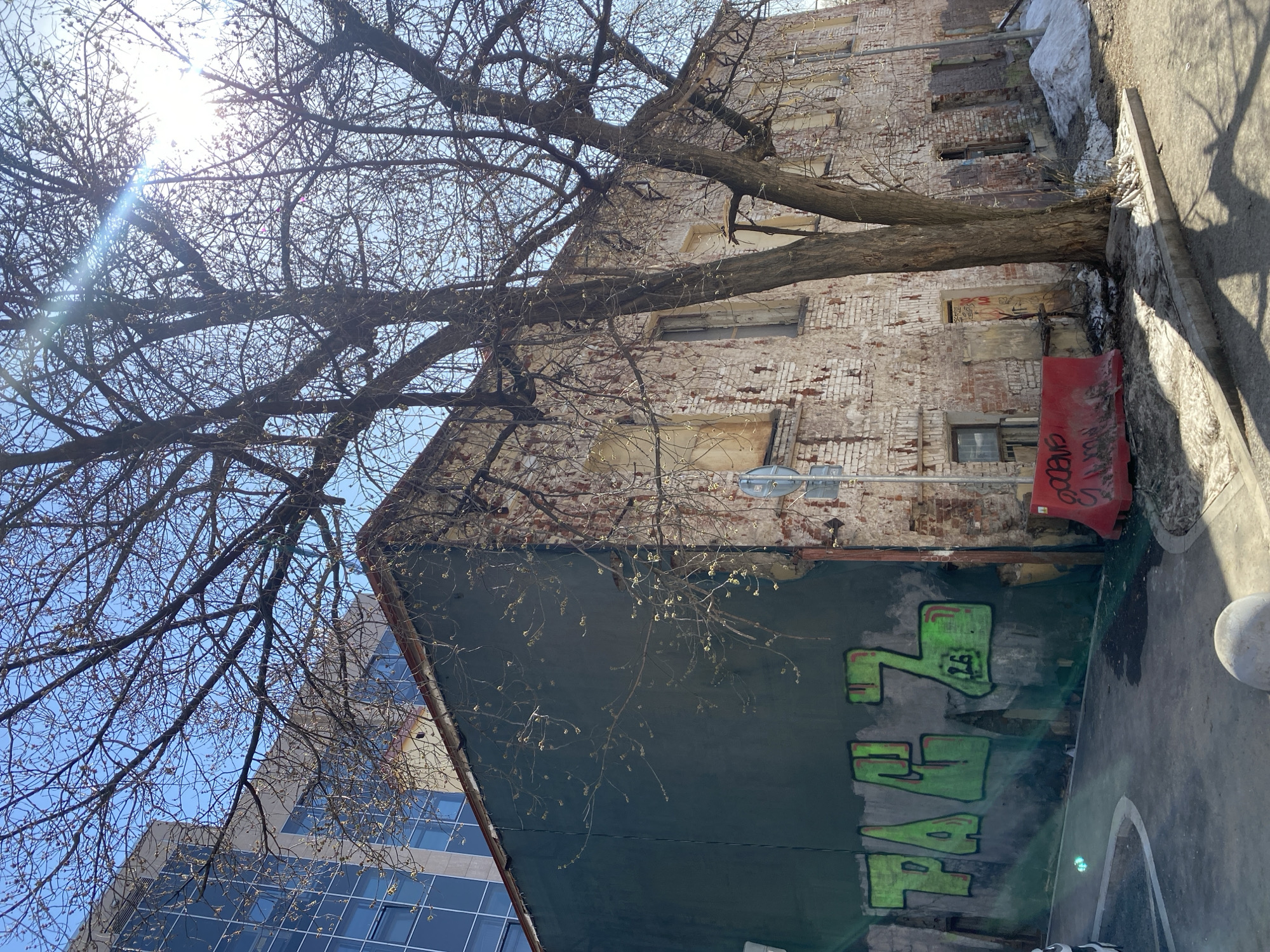 A ground-level, sunlit shot, taken outdoors, depicts a building at an angle with trees and a modern skyscraper visible in the background. The building in focus has a painted facade with graffiti in green, partially obscured by the trees. Above the painted section, the building's brick is visible. A tree with bare branches extends across the frame, obscuring parts of the building and sky. The modern skyscraper's windows reflect the sky. To the right of the building, there is a sidewalk. This image was taken in Moscow, Russia.