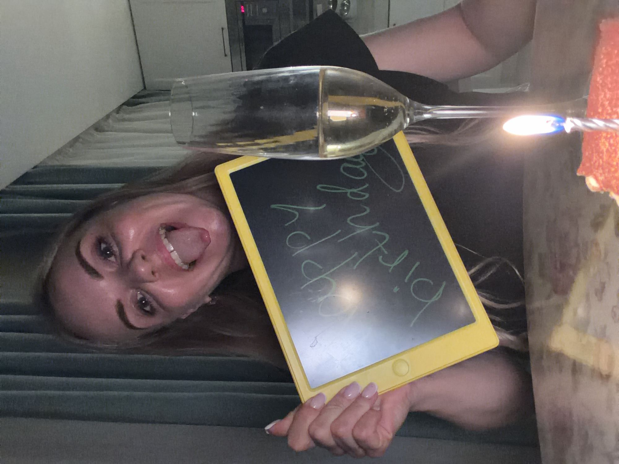 A woman is holding a tablet that reads "Happy birthday" next to a glass of champagne, a cake with a lit candle, and sticking her tongue out at the camera. The scene is indoors, and the background includes curtains and a kitchen area. There is no evidence in the image to indicate that the location is Votkinsk, Russia.