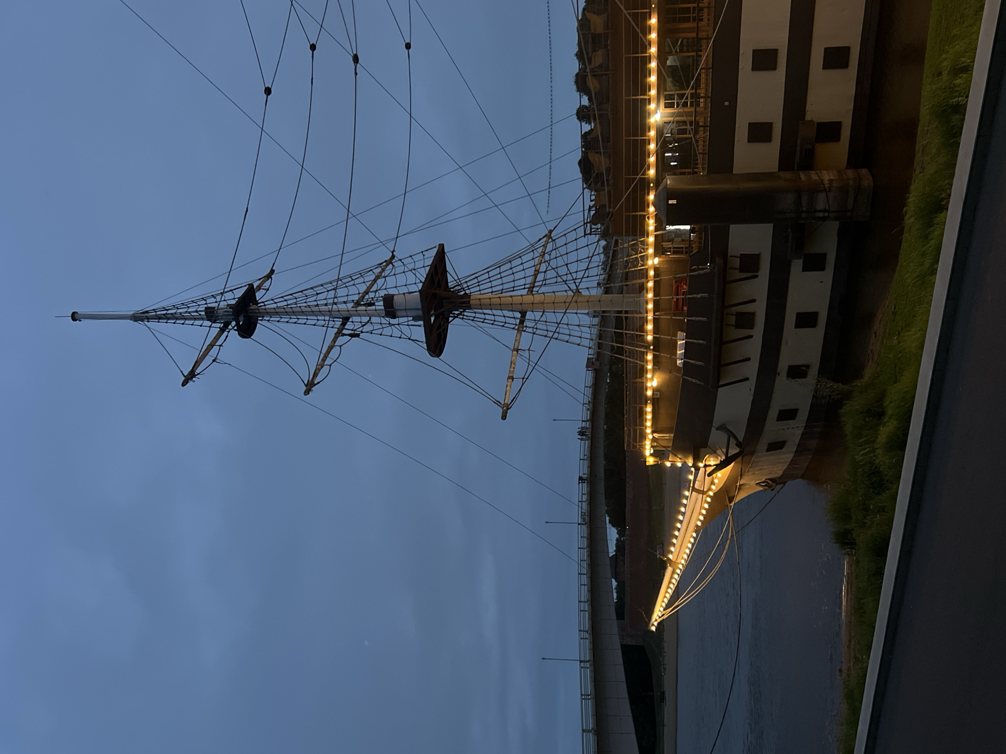 The image shows a large, old-fashioned sailing ship docked at night in the city of Veliky Novgorod, Russia. The ship has multiple masts and rigging, and its hull is painted in dark and light colors. String lights are illuminating the deck and the bow of the ship. In the background, a bridge spans a body of water, and a few people can be seen walking on it. The sky is dark and cloudy, suggesting it is evening or night. The ship is positioned along a grassy embankment.