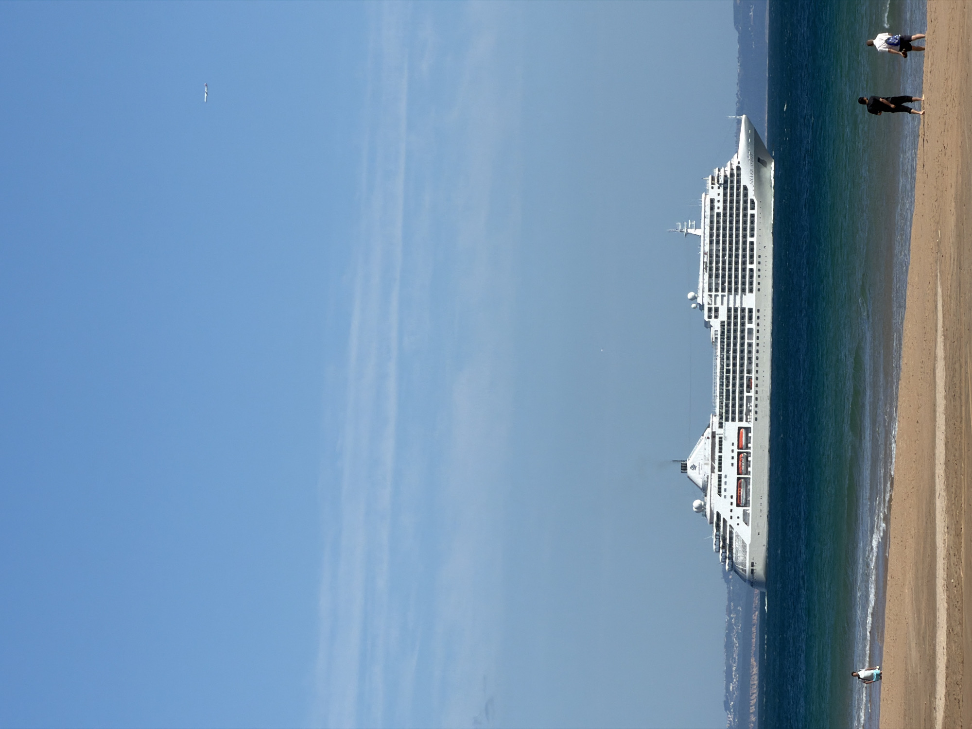 The image depicts a beach scene near Cascais, Portugal. A large cruise ship is anchored close to the shore. Several people are walking along the beach near the shoreline. The sky is clear with some wispy clouds.