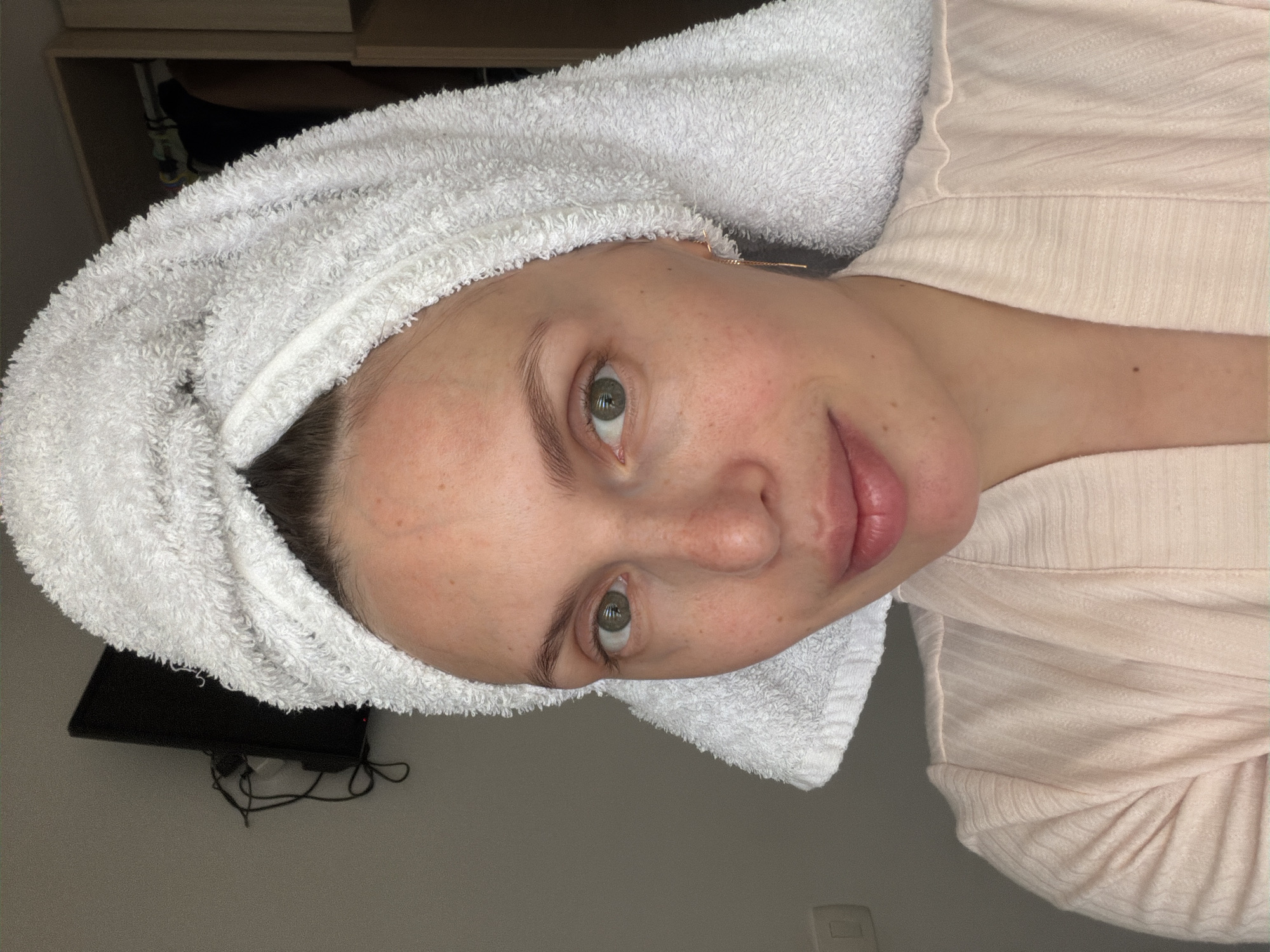 A person with fair skin, green eyes, and light brown eyebrows is centered in the image. She has a white towel wrapped around her head. The person is wearing a light pink or beige shirt. In the background, there is a cabinet, a television, and a wall. There is no information in the provided picture to determine if the location is in Budva, Montenegro.
