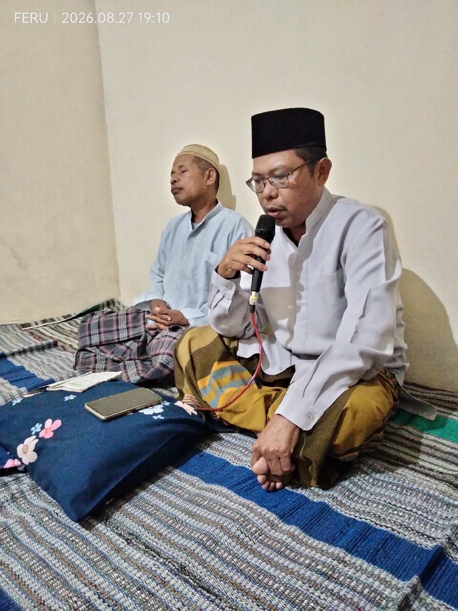 Two men are sitting cross-legged on a woven mat. The man on the right, wearing a black peci (skullcap) and glasses, is speaking into a microphone. He is wearing a light-colored, long-sleeved shirt and a sarong with yellow and orange stripes. His hands are clasped in front of him. The man on the left is wearing a light-colored cap and a light-blue shirt, also with a sarong that has plaid patterns. His eyes are closed, and his hands are resting in his lap. A dark blue pillow with floral patterns, a mobile phone, and what appears to be a thin booklet are placed on the mat in front of the men. The background is a plain, light-colored wall. The timestamp in the top left corner indicates the image was taken on August 27, 2026, at 19:10, and is tagged with "FERU". The scene appears to be indoors, possibly in a home or community gathering space in Boreng, Indonesia.