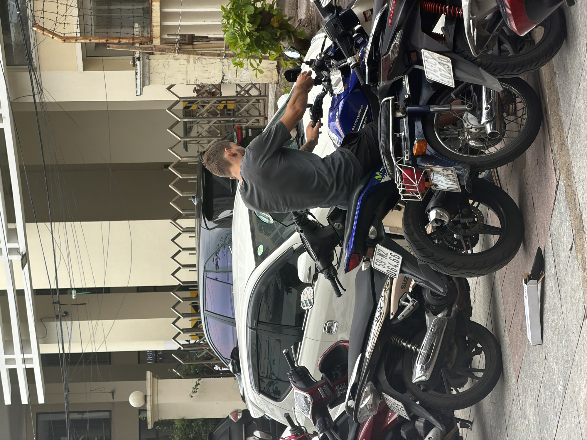 The image depicts a daytime street scene in Bac Nha Trang Ward, Vietnam, with clear weather. A man, wearing a grey t-shirt and dark shorts, is seated on a blue and black motorcycle, actively handling its handlebars. He appears focused, possibly preparing to ride or making an adjustment.
The man is positioned in a densely packed row of parked vehicles. A white car, possibly an SUV or minivan, is parked directly behind the man and his motorcycle. Multiple other motorcycles and scooters are parked closely together in the foreground and extending into the background, highlighting a common mode of transport in the region.
The setting includes light-colored multi-story buildings in the background, suggesting an urban or residential area. Green foliage from a plant is visible near one of the buildings. Overhead utility wires traverse the upper part of the scene. The ground is a light gray concrete or tiled sidewalk.
Visible text and details include:
- On the blue motorcycle: "bromb0" (likely Brembo), "Thalar", and a "Movistar" logo. Its license plate displays "59-A1 010.53".
- On a beige/goldish motorcycle further to the right: "SUZ" (Suzuki) branding and a license plate reading "59-M2 064.66".
- Another partially visible license plate in the background shows "40740".
- A white rectangular box lies on the sidewalk, with the text "INGH" visible on its side.