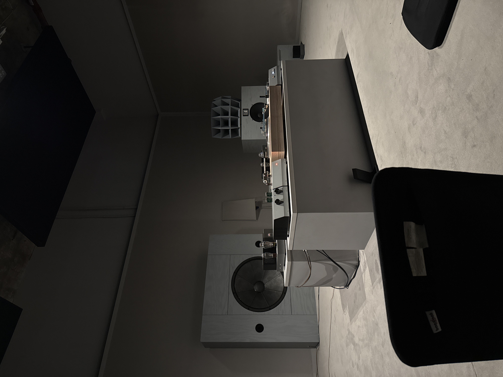 The image displays an interior view of a high-fidelity audio listening room or showroom, characterized by a minimalist aesthetic. The space features light grey walls and a matching light-toned floor, creating a subdued backdrop for the audio equipment. The lighting is dim, suggesting an evening setting or an intentionally low-lit environment designed for focused listening.
The central focus is a sophisticated audio system arranged on a long, light-colored console or cabinet. On the left side of the console, several electronic components are visible, including what appear to be vacuum tube amplifiers, which emit a subtle, warm glow. Other silver-toned audio devices with various controls are also present.
Two large, custom-designed speakers frame the console. The speaker positioned prominently in the foreground on the left features a large, dark central woofer and a small circular port, enclosed within a light-colored cabinet. Further back on the right, another speaker showcases a distinctive, multi-celled horn array at its top, indicating a design focused on precise acoustic dispersion.
Partial views of two black chairs are visible, positioned for listening. On a small tag attached to the back of the closest chair, the text "boo boo" is discernible. Cables are neatly managed behind and around the audio components. A dark, rectangular panel or screen is visible on the far left wall, and a small vent can be seen on the wall above the foreground speaker. The overall scene suggests a dedicated and refined space for an immersive audio experience in London, United Kingdom.