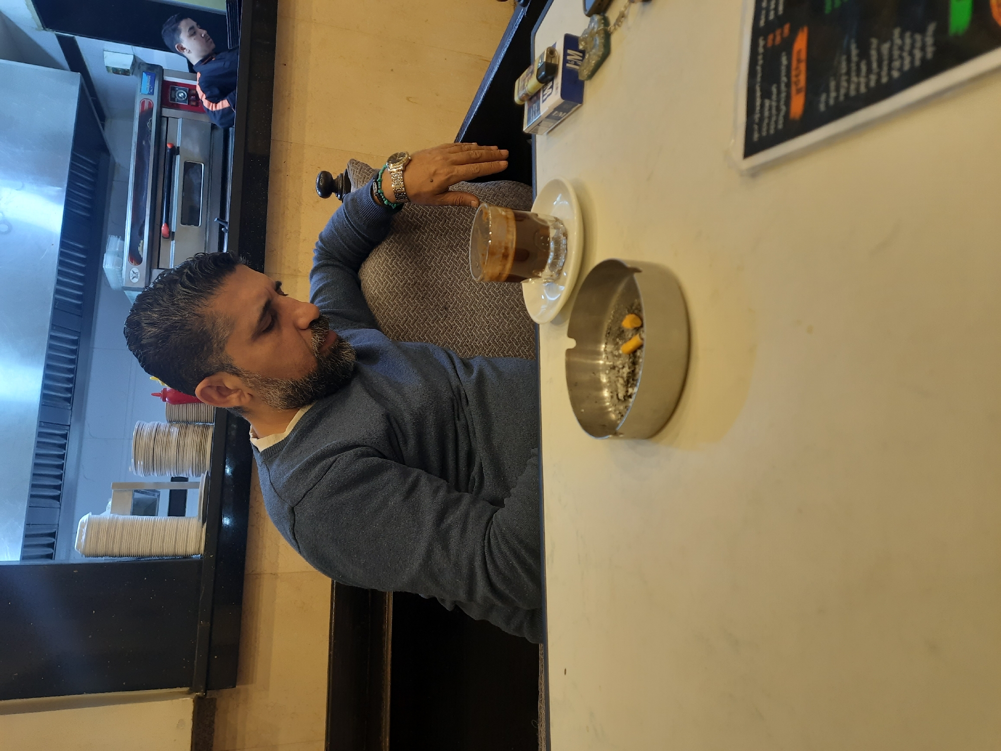 A man with dark hair and a beard is seated at a table in a restaurant, possibly in Unknown, Unknown. He is wearing a blue sweater, a watch, and a bracelet. On the table are a glass of coffee, an ashtray with cigarette butts, a pack of cigarettes, and a menu. Another person is visible in the background near what appears to be a kitchen area with an oven. The restaurant setting includes what looks like a stack of paper cups.