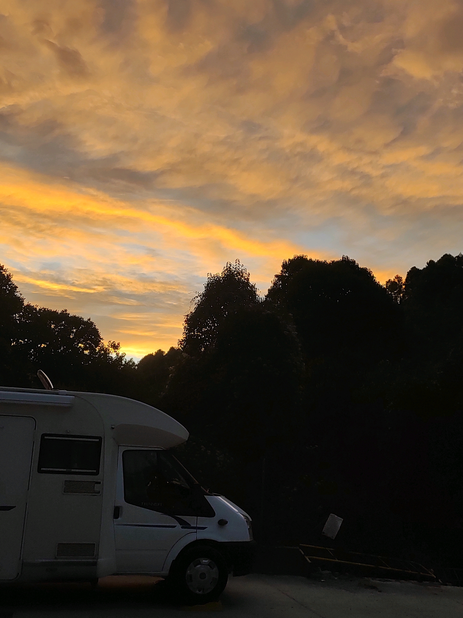 The image depicts an RV parked on a road in what appears to be a rural or semi-rural area, possibly at dusk or dawn given the warm, diffused light in the sky. The sky is filled with wispy clouds illuminated in shades of orange, yellow, and pale blue, suggesting a sunset or sunrise. Silhouetted against the sky are the dark outlines of trees, forming a dense treeline behind the RV.
The RV itself is white and appears to be a motorhome. It is parked on a paved surface, with the front of the vehicle visible on the right side of the frame. The side of the RV, facing the camera, shows windows and ventilation grates. The overall impression is one of a peaceful, perhaps solitary, scene in nature.
There are no people visible in the image. The setting is an outdoor environment with a focus on the RV and the natural backdrop. The lighting conditions, with the dramatic sky and silhouetted trees, create a serene and atmospheric mood. The specific location in Suera, Spain, is not directly evident from visual cues within the image itself, but the description assumes this context.