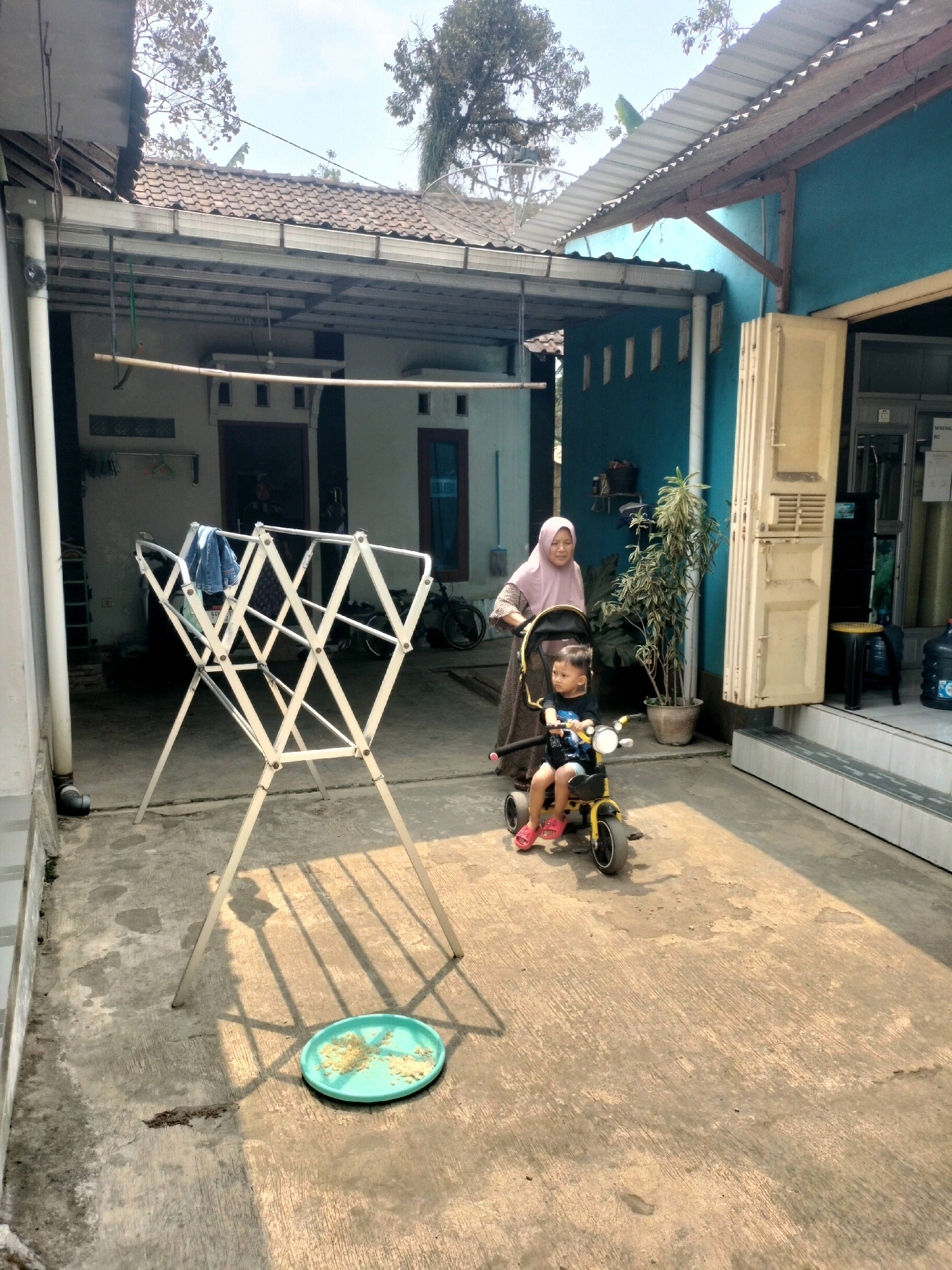 In Pujut, Indonesia, a middle-aged woman wearing a pink hijab and a patterned dress pushes a stroller containing a toddler. The toddler, dressed in a blue shirt and shorts, is riding a yellow tricycle. The scene takes place in an outdoor courtyard with a concrete floor and a clothes drying rack in the foreground. In the background, there is a house with a tiled roof and blue walls, along with a potted plant and some bicycles. The sun casts long shadows across the courtyard.