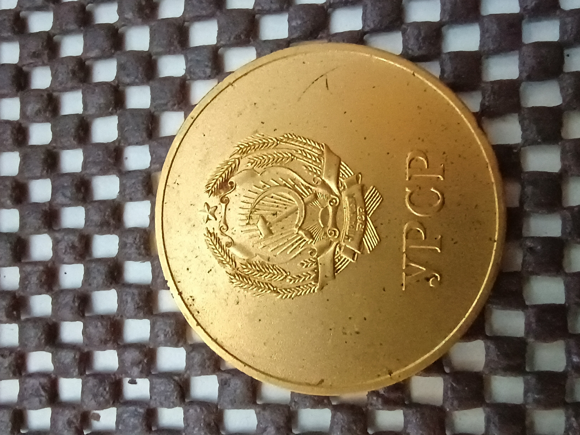 A close-up, overhead shot shows a gold-colored coin with embossed imagery resting on a textured, dark gray and white checkered surface. The coin features a central emblem depicting a hammer and sickle crossed with a star, surrounded by wheat stalks. Below the emblem, the Cyrillic letters "УРСР" are visible. The checkered surface consists of numerous small, raised, cube-like elements arranged in an alternating pattern, creating an open grid. The lighting is direct, casting subtle shadows. The image is sharp and focuses on the coin and its immediate surroundings. No people or other objects are present in the frame. The location in the city of Zaporizhzhia, Ukraine, is not discernible from the visual information provided in the image.