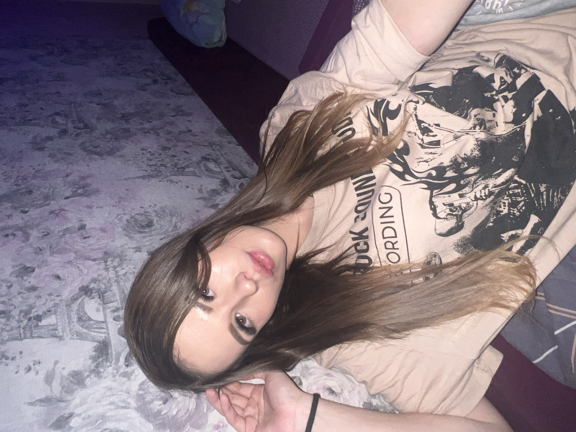 A young woman with long brown hair is lying on a floral-patterned surface, possibly a bed, adjacent to a dark purple cushion. She is wearing a beige t-shirt with a black graphic design that includes the word "recording." Her right hand is visible near the bottom of the frame, and she is looking towards the camera. The lighting is dim, and the orientation of the image is tilted. The location information of Noviy, Russia, is not discernible from the visual data.
