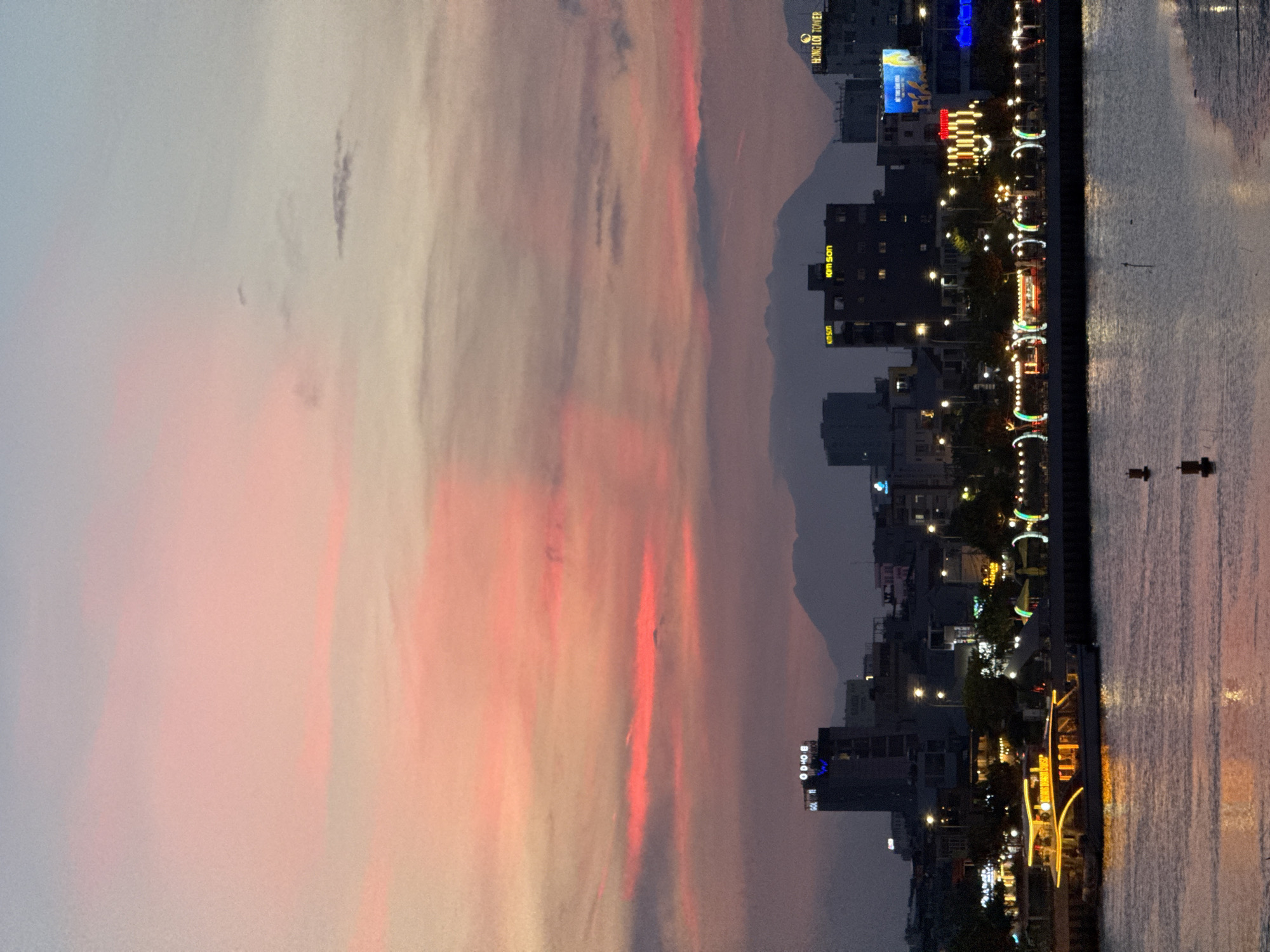 The image depicts a waterfront scene in Đà Nẵng, Vietnam, at dusk. The sky is a gradient of soft pinks and oranges from the setting sun, transitioning to a pale blue overhead with some wispy clouds. In the distance, silhouetted mountains form a backdrop. The city skyline is illuminated, featuring several buildings of varying heights. Prominent among these are buildings with illuminated signage, including "HỒNG LỘI TOWER" and "KIM SON". The waterfront promenade is lined with trees and decorated with string lights and festive light displays. A large, illuminated tour boat with a yellow glow is docked or moving slowly on the calm water in the foreground. The water reflects the city lights and the colors of the sky. There are no visible people or specific actions occurring, creating a serene and ambient atmosphere of a city winding down for the evening.