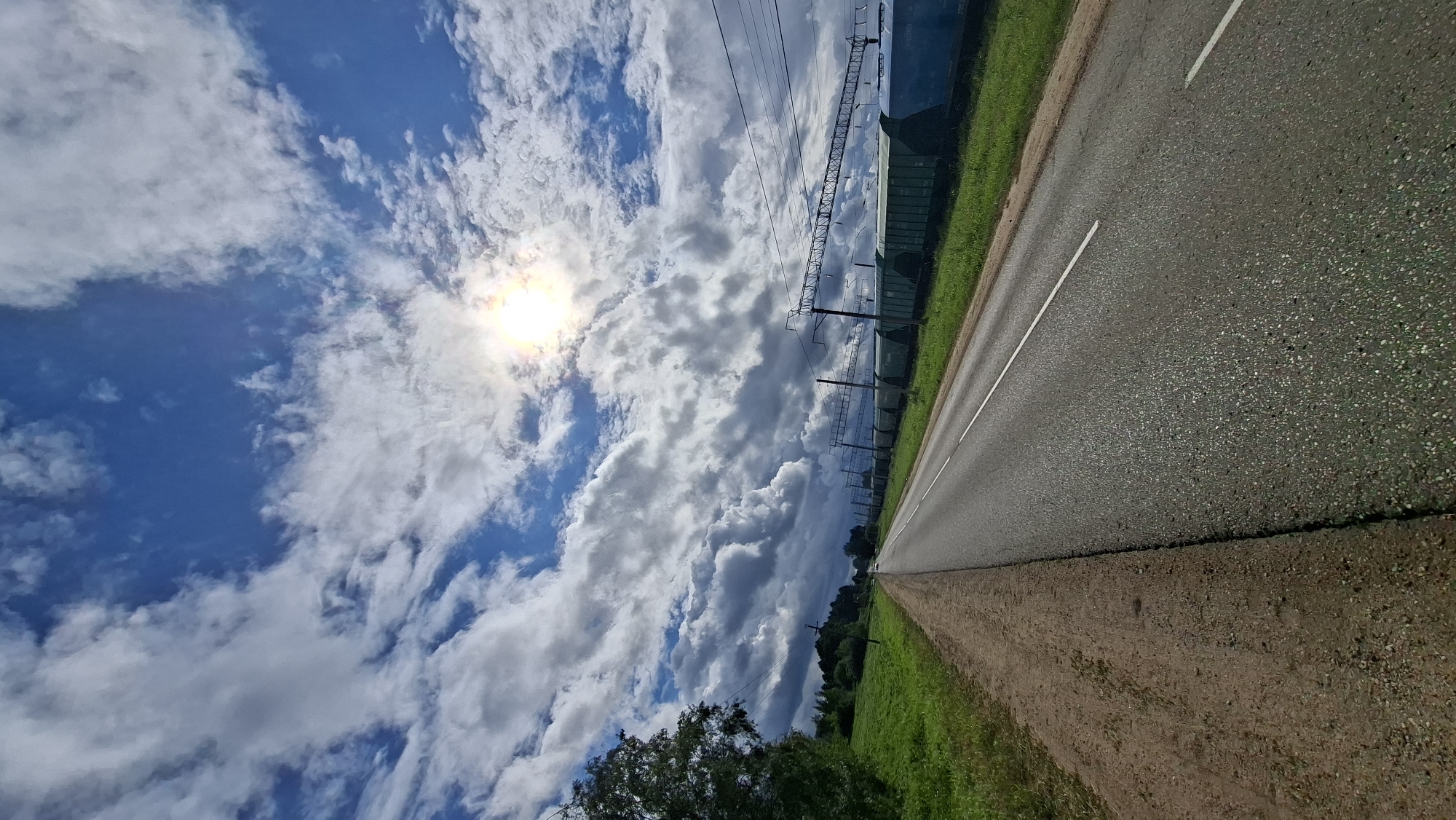 The image captures a rural roadside scene in the vicinity of Smyalyavichy, Belarus, under a partly cloudy sky with the sun partially obscured by clouds. A paved asphalt road with white lane markings stretches into the distance, bordered on the left by a dirt shoulder and grassy fields. On the right side of the road, a long train composed of green and blue freight cars is stationary, with overhead power lines supported by metal pylons running parallel to the tracks. A few trees are visible on the far left of the frame, with more distant foliage suggesting a wooded area. The overall atmosphere is that of a bright, albeit slightly overcast, day in a quiet, possibly industrial or transport-hub-adjacent rural area. There are no people visible in the image.