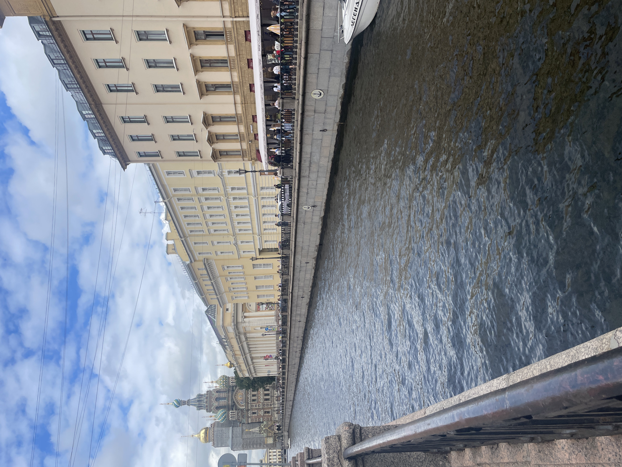 This is a wide-angle, eye-level, outdoor photograph of a canal in Saint Petersburg, Russia. The canal is the dominant element, occupying the lower half of the frame. The water is dark blue with ripples and reflections. Along the right bank of the canal are several buildings with light-colored facades and multiple windows. The buildings appear to be of historical architecture, with some displaying ornate detailing. A canopy, likely from an outdoor cafe or restaurant, stretches along the base of these buildings. Several people are visible standing or sitting along the canal's edge, some near the cafe. A white boat with a dark stripe is docked to the right of the frame, with some text visible on its side.
On the left side of the canal, a distinct church with multiple golden domes and intricately decorated facades stands out against the sky. This is likely the Church of the Savior on Spilled Blood, a famous landmark in Saint Petersburg. Further to the left, a stone embankment with a dark railing lines the canal. The sky above is partly cloudy with scattered white clouds against a blue background. Power lines are visible strung across the upper portion of the image. The overall scene depicts a busy urban waterway, characteristic of Saint Petersburg's many canals and historical architecture.