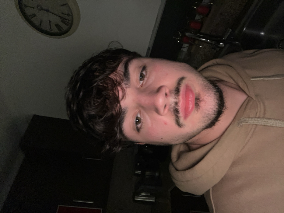 A selfie captures a young man with dark, wavy hair and a trimmed beard, wearing a tan hoodie. He gazes into the camera with light eyes and a slightly parted mouth. The background features a clock on the wall, dark cabinets, and a glimpse of a kitchen sink area with metallic fixtures. The lighting is dim, casting shadows and emphasizing the man's features.