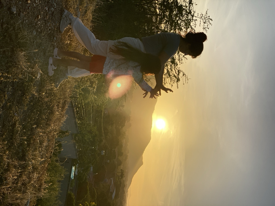 A woman wearing white pants and a grey shirt is bent over a child wearing red shorts and a white shirt. They are positioned in front of a sunset backdrop with a mountain silhouette. Their hands are outstretched toward the sun. The foreground includes a grassy terrain and a partial view of a fence-like structure. The overall scene is bathed in warm, golden light.