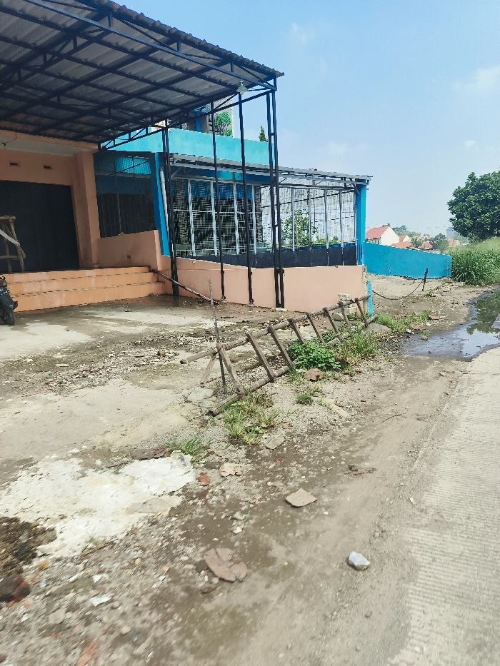The image shows a dirt road leading to a building with a blue wall and a metal roof. The building has a large window and is located in Cileunyi, Indonesia at latitude -6.947 and longitude 107.727. The ground is covered in dirt and debris. There is a wooden ladder leaning against the side of the building. There is no one visible in the image. A small, rusted, metal object can be seen in the lower right of the frame. The area appears to be under construction or renovation.