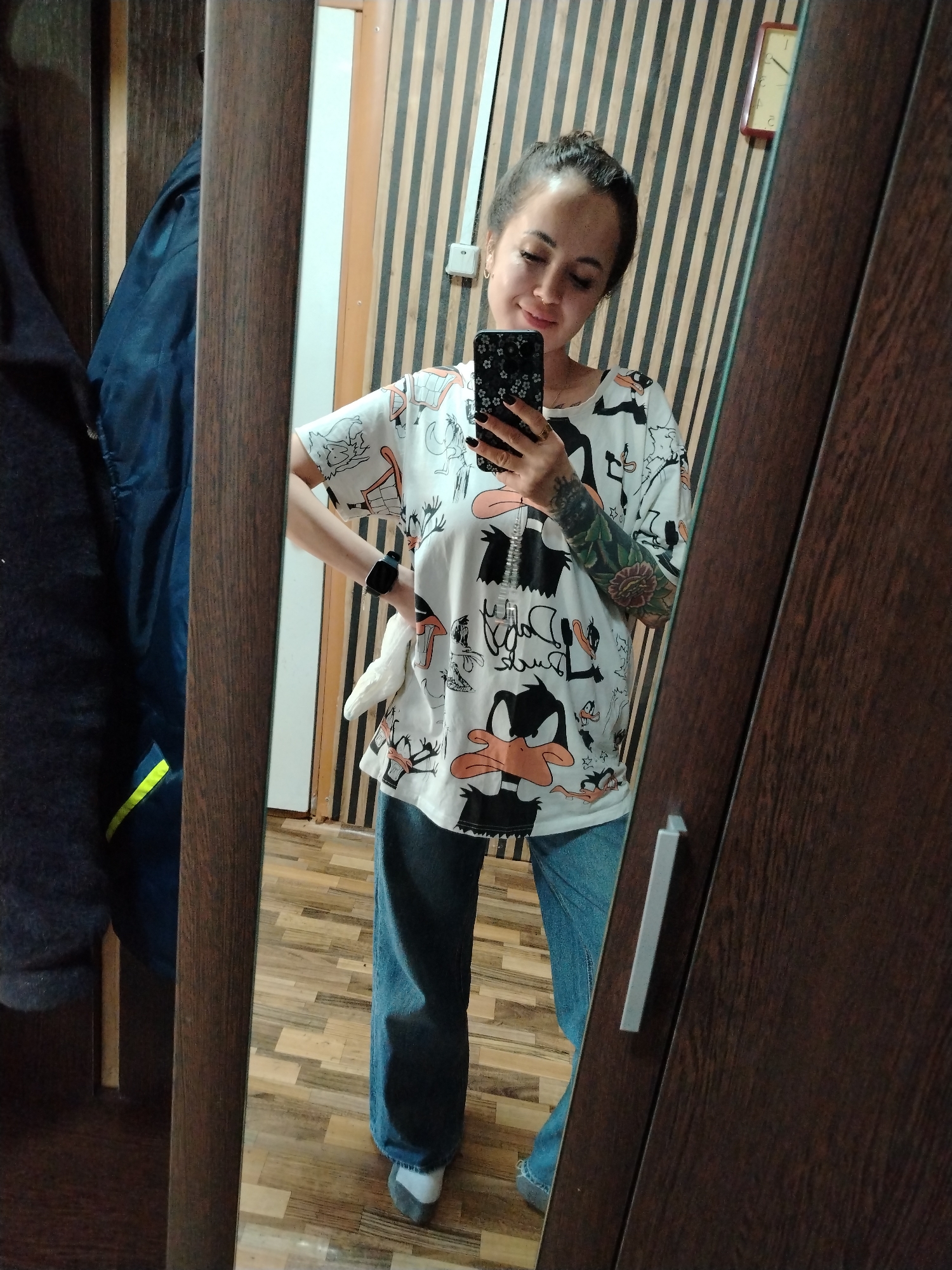A young woman is taking a selfie in a mirror. She has her hair pulled back in a bun and is wearing a white t-shirt with cartoon characters and blue jeans. Her left arm has a tattoo. She is holding a cell phone in her right hand. The mirror is attached to a brown wooden cabinet. Behind her, the wall is decorated with vertical stripes. A coat hangs on the left side of the cabinet. The location appears to be indoors. The clock visible on the wall suggests the location could be in Гожан, Russia, although this is not confirmed within the image itself.