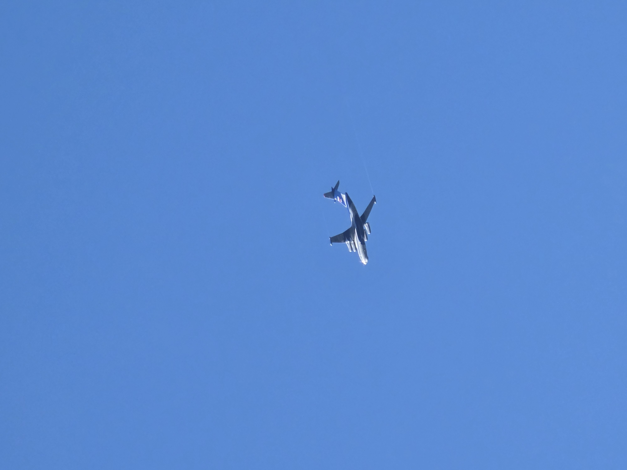 A large, grey and blue seaplane is captured in mid-flight against a clear blue sky. The aircraft appears to be a Beriev Be-200, identifiable by its distinctive high wing and T-tail configuration. A faint white contrail trails behind the plane. The image shows the seaplane from a side-rear perspective, with its engines mounted above the wings. The location is given as Речицы, Russia, although the image itself provides no visual cues to confirm or deny this specific location. The lighting suggests it is daytime, with the sun likely positioned to the right and slightly behind the viewer, illuminating the left side of the aircraft.