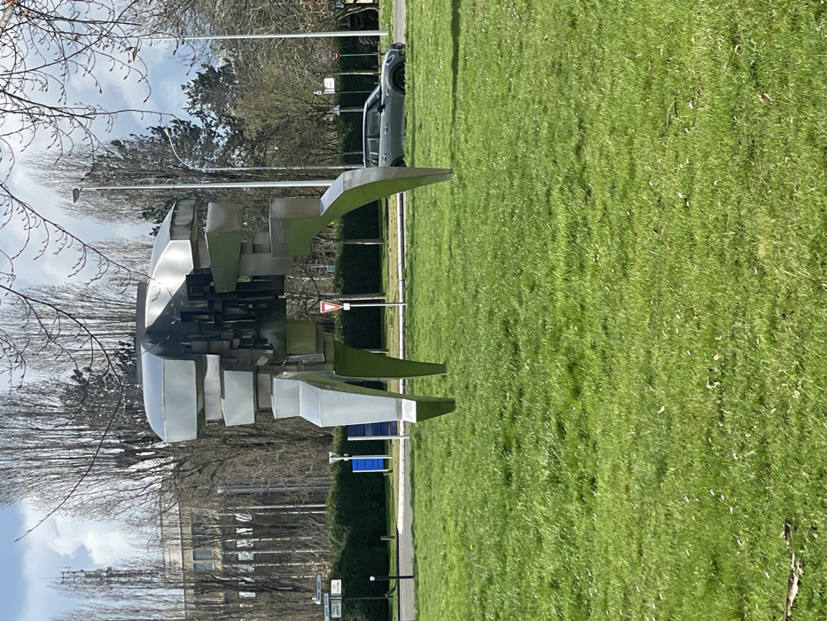 The image displays an outdoor urban environment in Montigny-le-Bretonneux, France, under natural daylight. A large, abstract, bipedal metallic sculpture dominates the central frame, positioned on a vibrant green lawn. The sculpture is constructed from angular, reflective stainless steel panels, forming a complex, segmented body with two prominent, splayed legs in the foreground and two in the background. The immediate foreground features bright green grass, flecked with small white or yellow flowers.
Behind the sculpture, a paved road or driveway is visible. A dark grey four-door Mini Countryman car is parked facing right on the roadside, partially obscured by a low dark green hedge and one of the sculpture's legs. To the left of the sculpture, a red-bordered white triangular "Cédez le passage" (Yield) road sign is present, alongside a blue rectangular sign and two streetlights. The background features a dense line of tall, bare-branched deciduous trees, suggesting late winter or early spring. Beyond these trees on the far left, a multi-story, light-colored building with visible windows stands, topped by a slender telecommunications tower. The sky above is light blue with scattered white clouds. No individuals are visible.