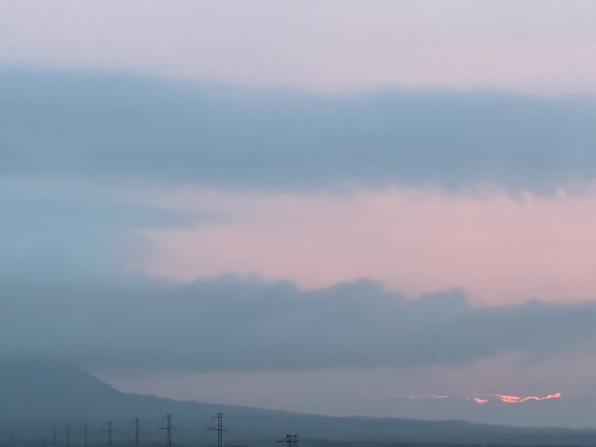 This image captures a twilight scene in Yuzhno-Kurilsk, Russia. The sky is filled with soft hues of blue and pink, indicating either dawn or dusk. Prominent horizontal bands of clouds stretch across the frame, with the lower band exhibiting a gentle, undulating texture. In the distance, a mountain range is visible, its peaks softened by the atmospheric conditions. A line of power pylons stretches across the foreground, silhouetted against the muted landscape. In the lower right quadrant of the image, a strip of cloud is illuminated with a vibrant pinkish-orange glow, suggesting the sun is either rising or setting just beyond the visible horizon, or peeking through a gap in the clouds. The overall impression is one of serene natural beauty, punctuated by human infrastructure.