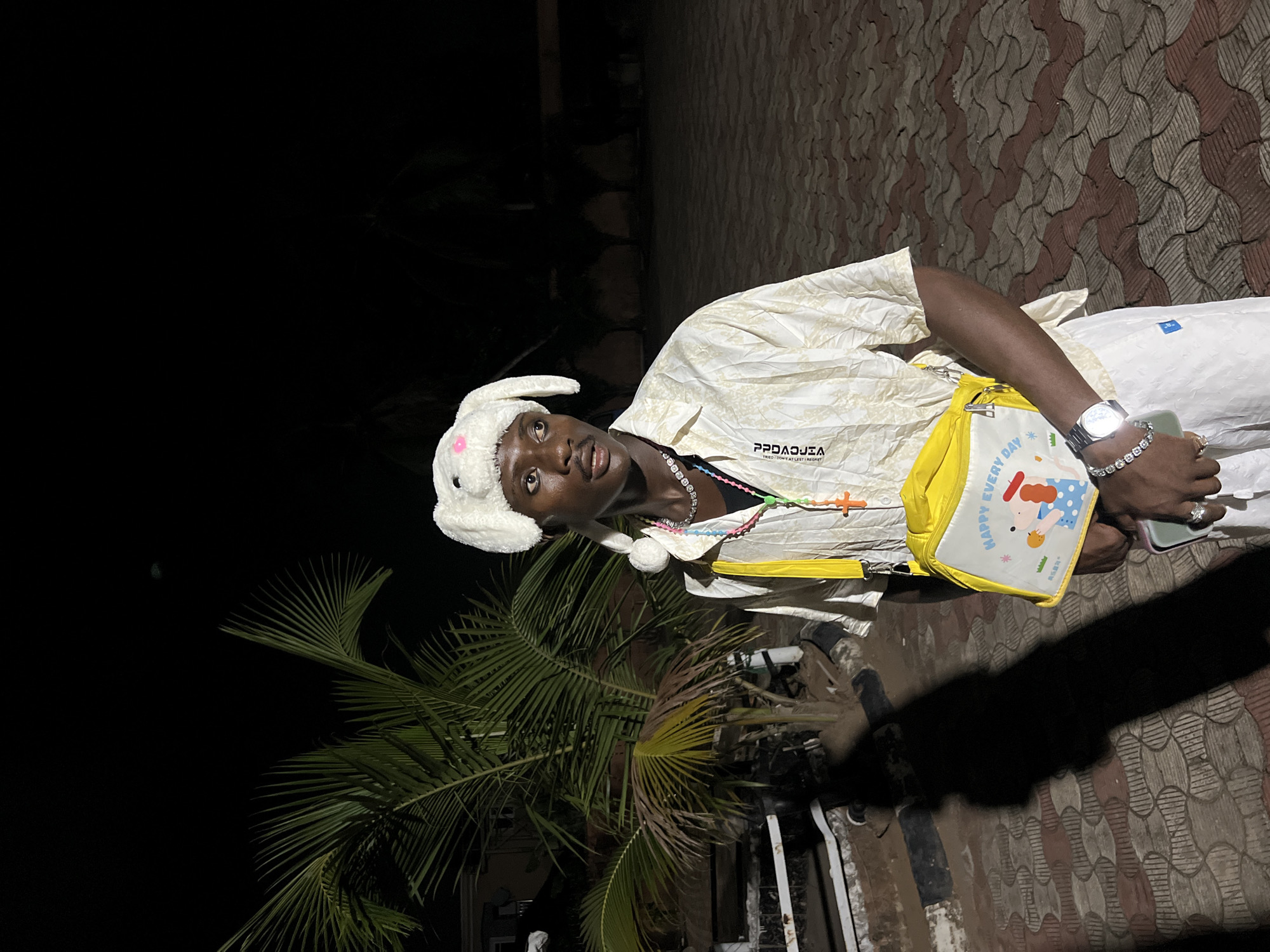 A young person stands outdoors at night in Unknown, Unknown. They wear a white bunny hat, a patterned beige shirt, white pants, and a yellow shoulder bag. They wear a silver watch and a diamond bracelet on their left wrist and hold a smartphone. A palm tree is visible in the background to the left, and the ground is paved with red and beige bricks.
