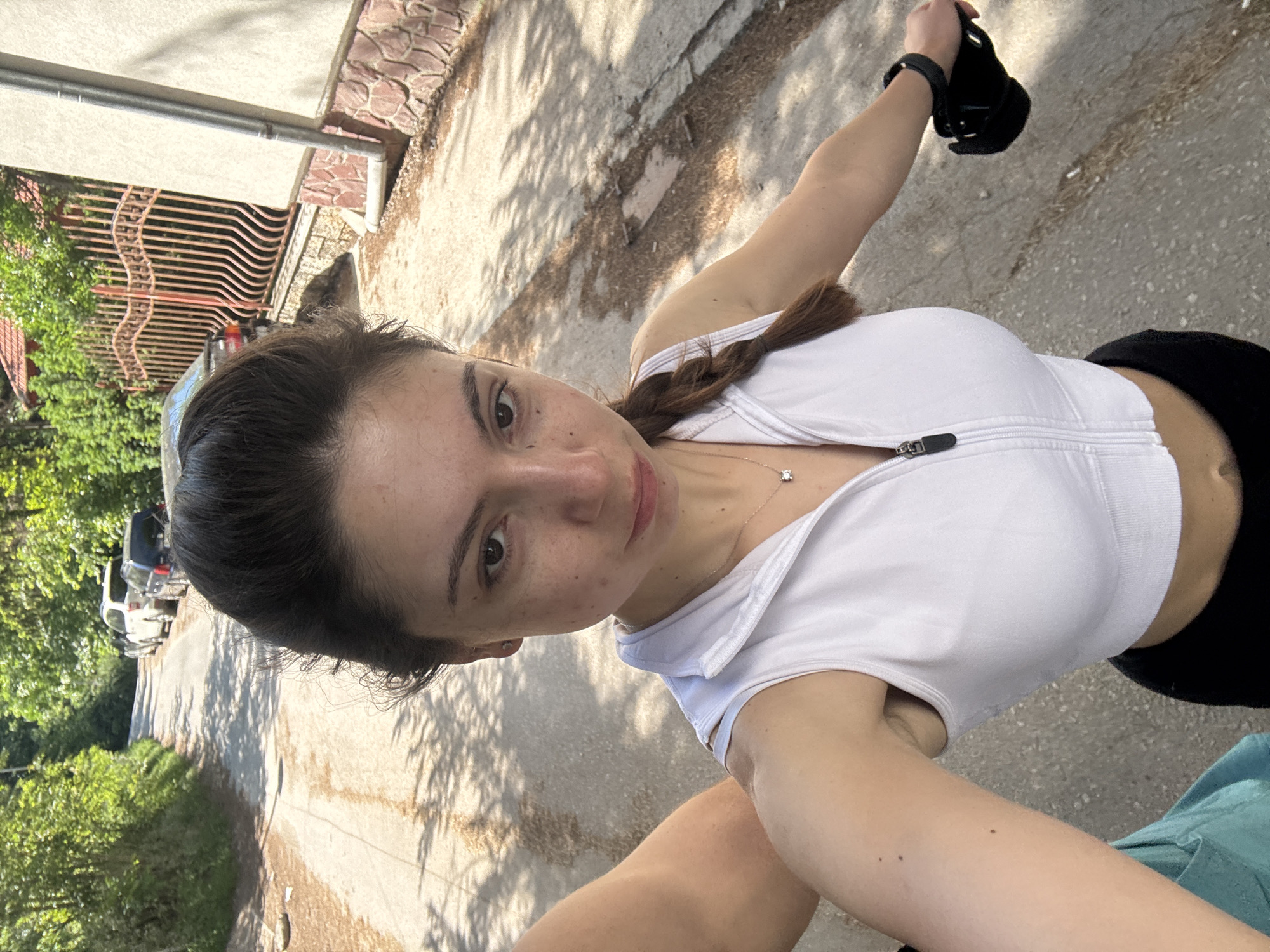 A young woman is taking a selfie outdoors. She is fair-skinned with dark hair in a braid and is wearing a white athletic zip-up top and black bottoms. She is holding a black object in her raised right hand. A silver necklace is visible around her neck. The background shows a paved road, a building with a red tile roof and wrought iron fence, green trees, and parked cars. The location appears to be a residential street, possibly within the city of Varna, Bulgaria, given the architectural style of the building. The environment is sunlit, casting shadows on the road.