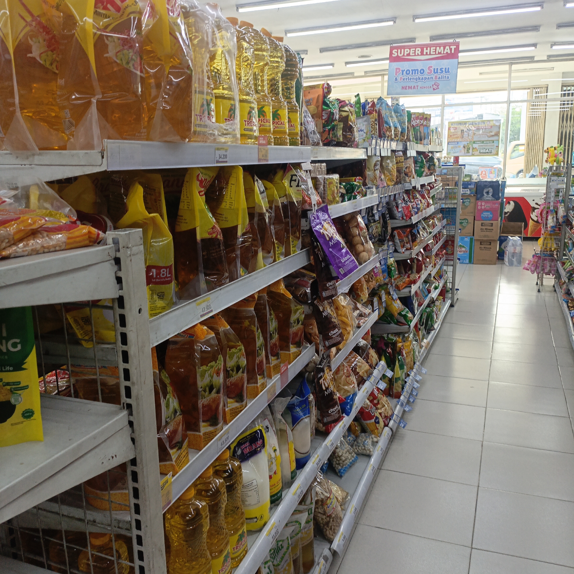 The image shows an aisle in a convenience store, possibly a 7-Eleven or similar establishment, in Salatiga, Indonesia. The shelves are stocked with various grocery items, including bottles of cooking oil, bags of snacks, and packaged food products. The aisle is well-lit by fluorescent lights. A promotional banner hangs from the ceiling, advertising "SUPER HEMAT Promo Susu & Perlengkapan Balita" which translates to "Super Save Milk & Baby Supplies Promotion". The floor is tiled with light-colored square tiles. No people are visible in the image. The overall impression is that of a typical retail environment.