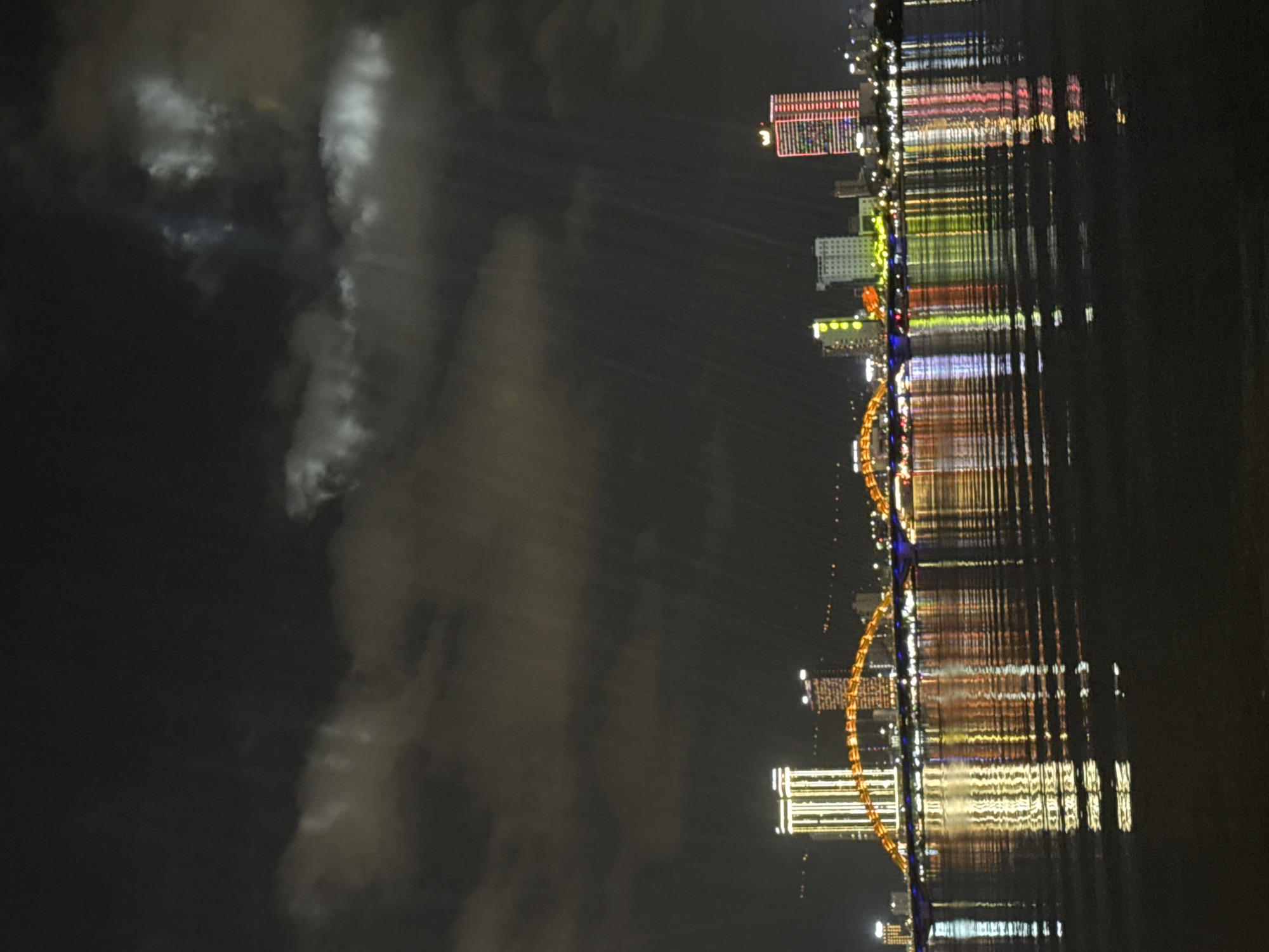 The image displays a captivating night view of a vibrant cityscape along a waterfront, reflecting dramatically on the water's surface. The scene is from Da Nang, Vietnam, likely featuring its iconic Dragon Bridge.
Numerous tall buildings line the riverbank, each adorned with a kaleidoscope of lights in red, green, yellow, and white, creating a brilliant urban spectacle. A distinctive arch bridge, characteristic of the Dragon Bridge, spans across the water, illuminated with bright yellow and orange lights that highlight its structural design. These colorful city and bridge lights produce long, shimmering reflections on the dark, slightly rippled water below, creating abstract vertical streaks of color.
The sky above is dark, but on the left side of the frame, dramatic, brightly illuminated clouds are visible. Streaks of light, appearing as beams or rays, cut through the sky and clouds, originating from the city. This illumination of the clouds and the presence of light beams suggest a festive atmosphere, possibly indicating a light show or other special event occurring in the city. No people or visible text are discernible in the image. The time of day is clearly night, and the weather appears to be cloudy, with the clouds themselves contributing to the visual drama due to the city's strong lighting.