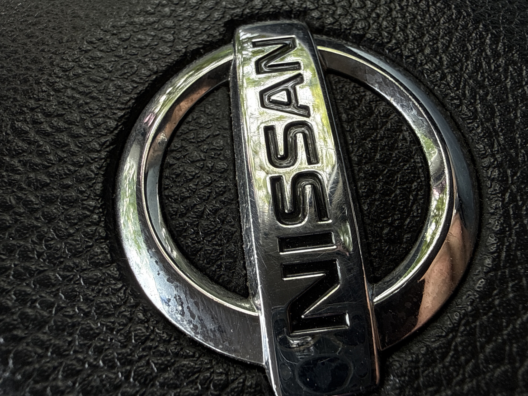 The image provides a close-up, high-angle shot of a metallic Nissan logo set against a dark, textured background. The central subject is the chrome emblem, featuring the word "NISSAN" in raised, uppercase letters on a horizontal bar that passes through the center of a circular chrome frame.
The logo's chrome surface is highly reflective, capturing bright, distorted reflections of its surroundings, including what appears to be green foliage or a bright sky, suggesting an outdoor setting during daylight. Evidence of wear is visible on the logo, including minor scratches and some subtle discoloration or patina on the chrome, particularly along the edges of the circle and within some letter recesses, showing a faint reddish-brown tint in places.
The background is a deep black, textured material, likely the surface of a car interior component such as a steering wheel or dashboard. Its granular, slightly irregular pattern provides a tactile contrast to the smooth, reflective metal of the logo. There is no specific activity depicted, as the image is a static shot of the emblem. While the location context is Vladivostok, Russia, this close-up image does not offer any direct visual cues to identify the city. The focus is entirely on the brand mark and its immediate material context.
