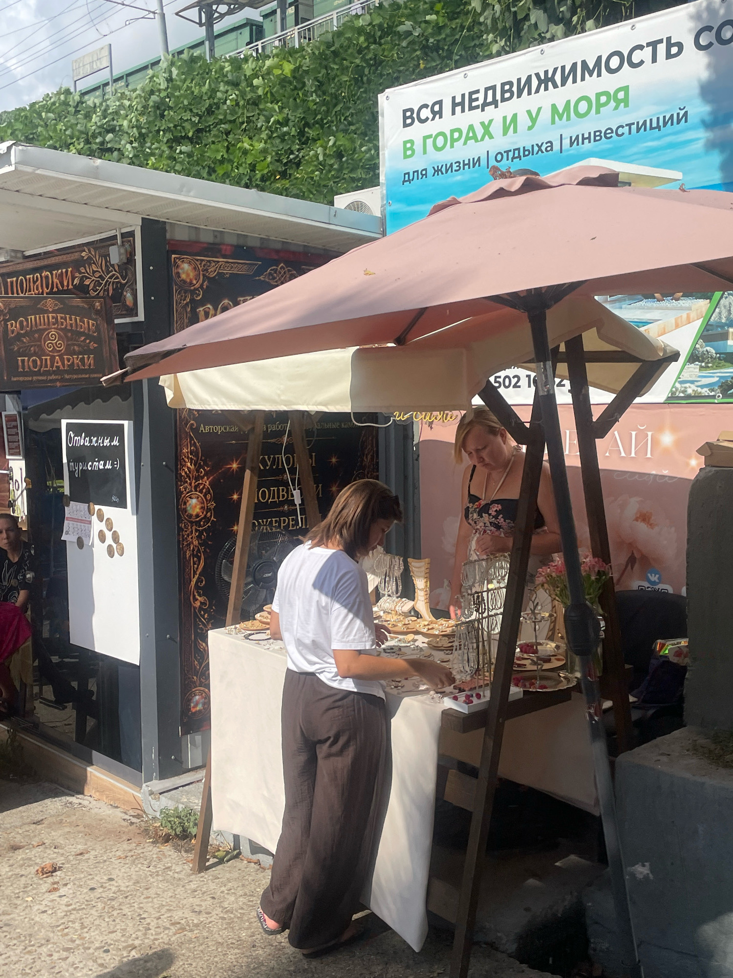 A full shot captures a bustling outdoor market stall in Sochi, Russia, likely on a sunny day. The stall is adorned with a pink and cream striped awning, offering shade to the displayed merchandise. A woman in a white t-shirt and dark, wide-legged pants is busy arranging jewelry on a table covered with a white cloth. She is facing away from the camera, her hair down, and her focus is on the items before her. Next to her, another woman in a floral-print dress stands, also attending to the stall.
Behind the stall, a large banner advertises real estate with text in Russian, including "ВСЯ НЕДВИЖИМОСТЬ СО" (ALL REAL ESTATE), "В ГОРАХ И У МОРЯ" (IN THE MOUNTAINS AND BY THE SEA), and "для жизни | отдыха | инвестиций" (for living | vacation | investment). The banner also includes a phone number and what appears to be social media icons.
To the left of the stall, a blackboard displays "Отважным туристам =)" which translates to "To brave tourists =)". Below this text, several circular stickers are affixed to the board. Various signs with decorative lettering and ornate designs are visible on the stall's facade, one of which reads "ПОДАРКИ" (GIFTS) and another "ВОЛШЕБНЫЕ ПОДАРКИ" (MAGIC GIFTS). The ground in front of the stall is paved with pebbles, and patches of green vegetation are visible near the stall's base. A metallic fan is positioned behind the table. In the background, lush green foliage covers an elevated structure, possibly a railway embankment. A woman wearing a dark top and pink scarf is partially visible on the far left, seated or leaning. The overall scene suggests a vibrant commercial area catering to tourists.